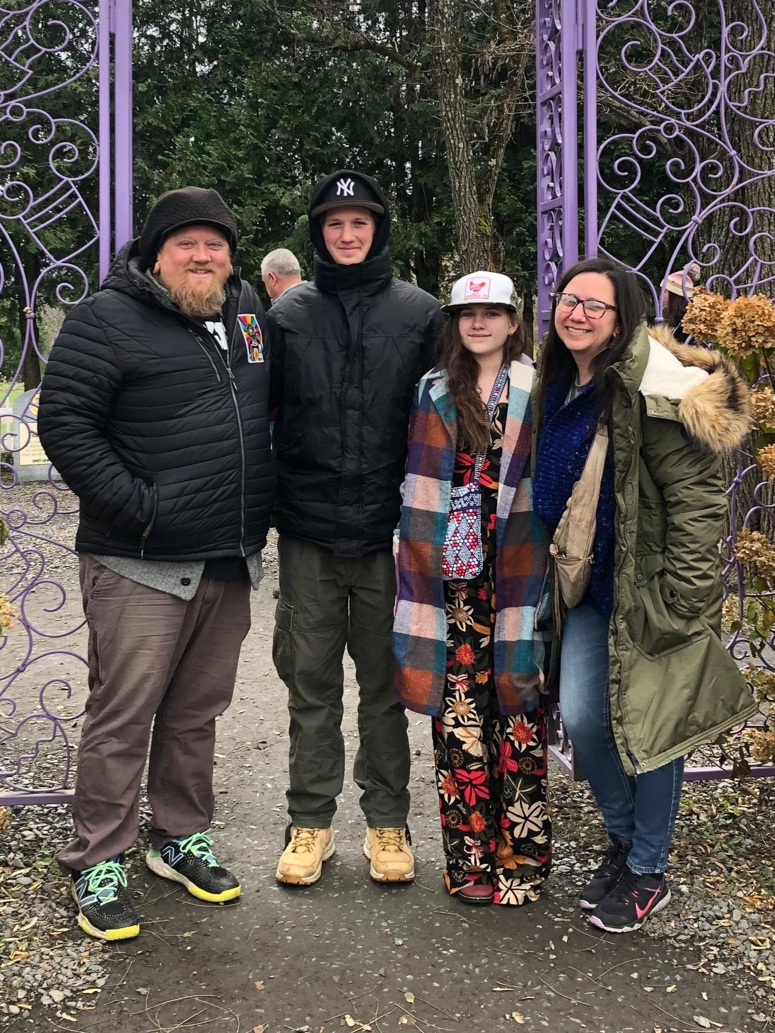 A family of four standing outdoors in front of a purple metal gate, smiling. The group includes two adults and two teenagers, dressed in casual winter clothing and jackets. The background has trees and a cloudy sky.