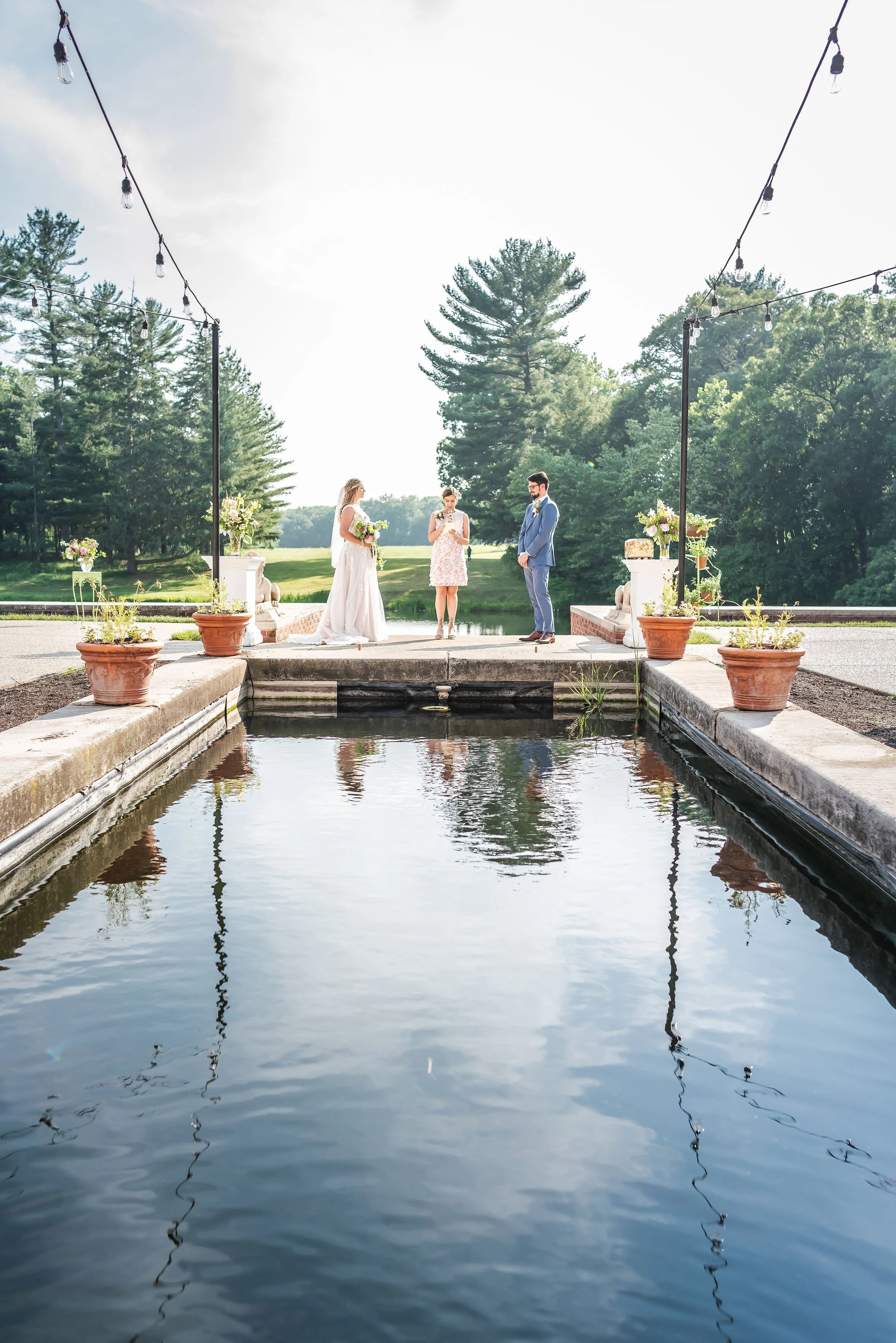 A wedding ceremony taking place outdoors by a pond, with trees in the background. The bride and groom stand facing each other with an officiant in between. The ceremony area is decorated with flowers and potted plants, and string lights hang overhead.