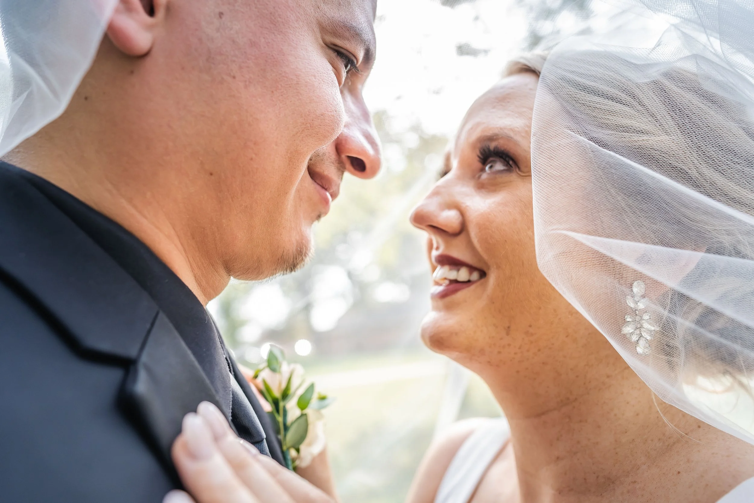 Close-up of a bride and groom looking into each other's eyes outdoors, with soft natural light behind them; the groom is dressed in a dark suit and the bride is smiling, wearing a wedding veil and jewelry.