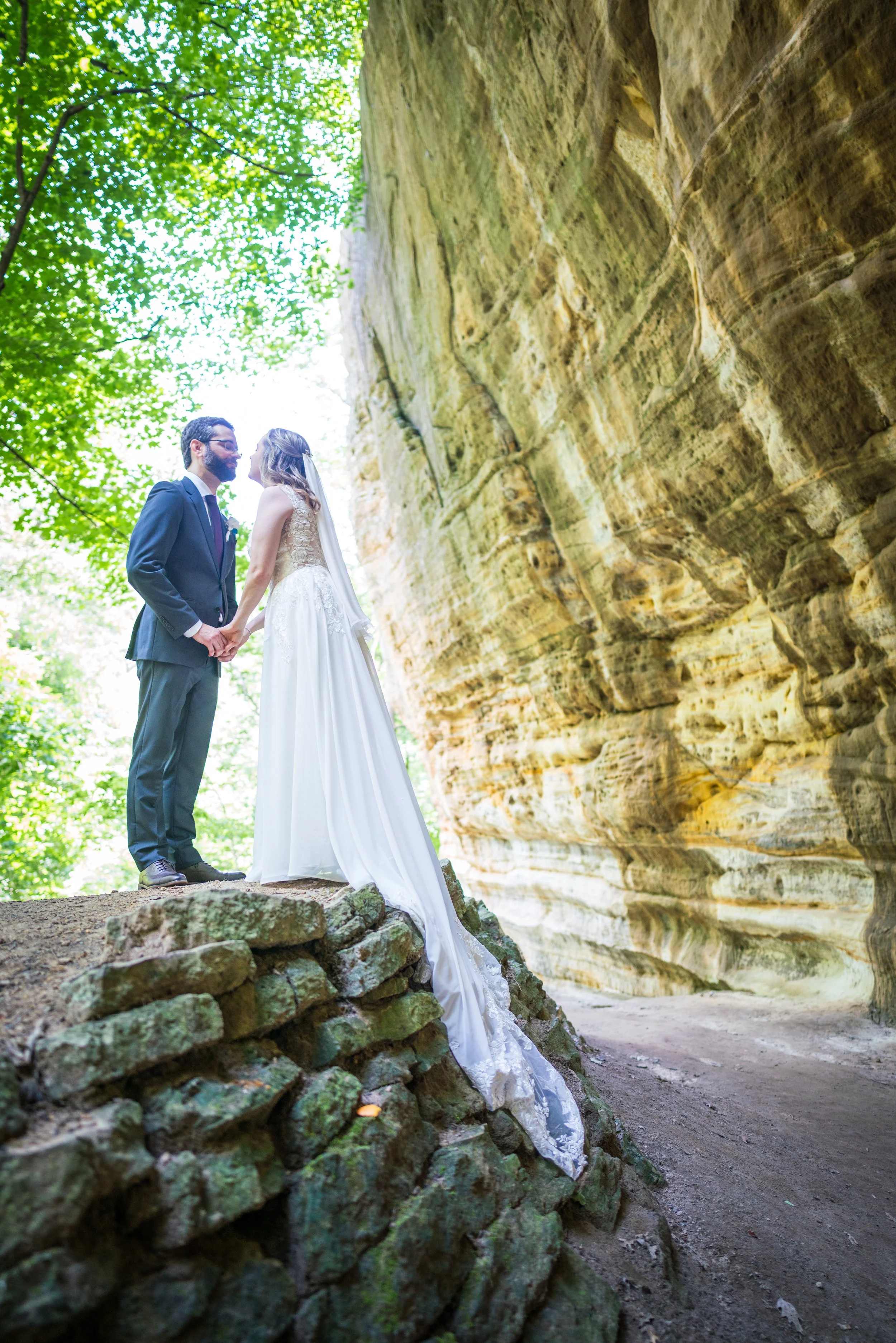 Bride and groom holding hands and standing on a rocky ledge outdoors, near a large rock formation and green trees, during their wedding ceremony.