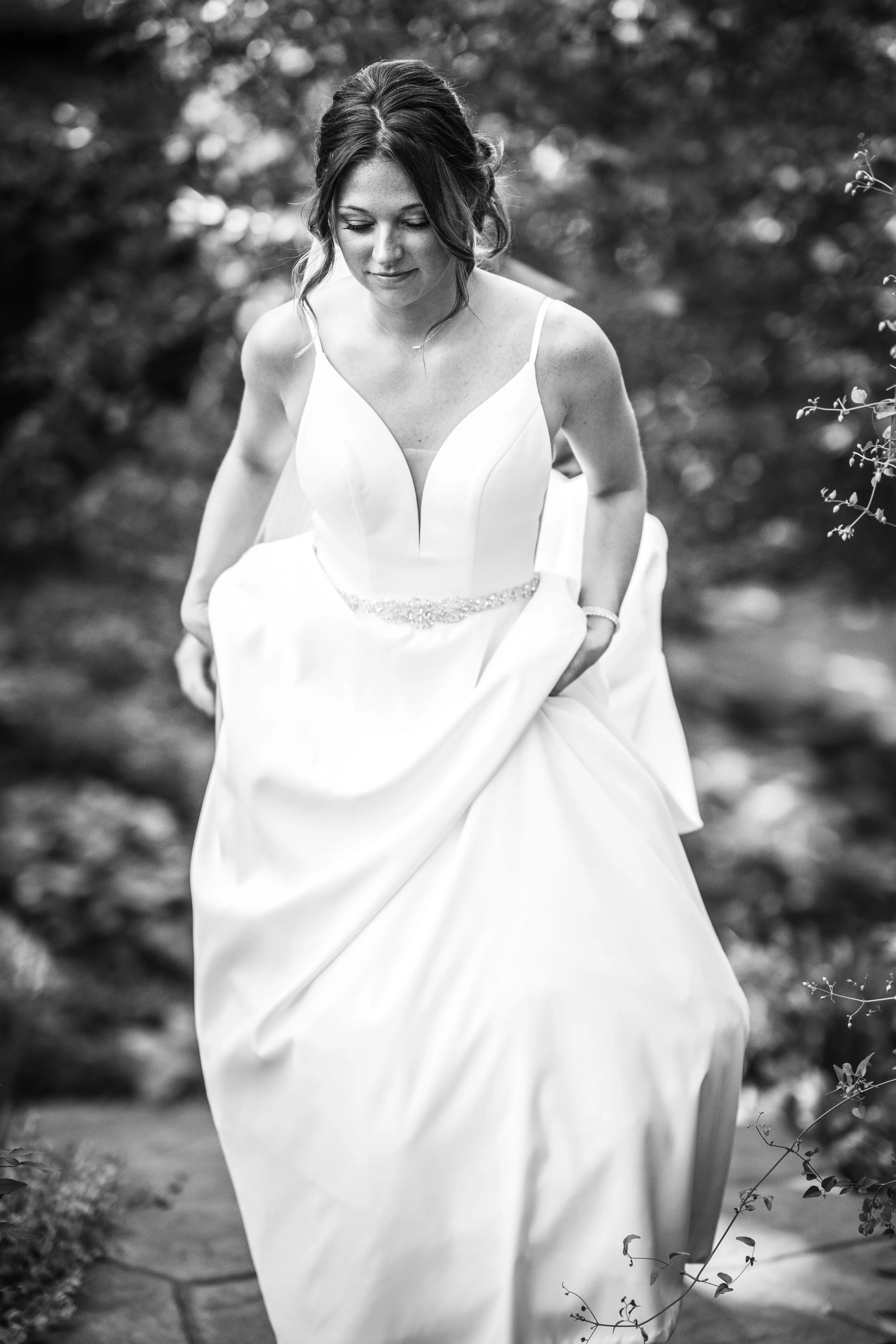 A woman in a wedding dress walking outdoors, looking down, with a blurred natural background.