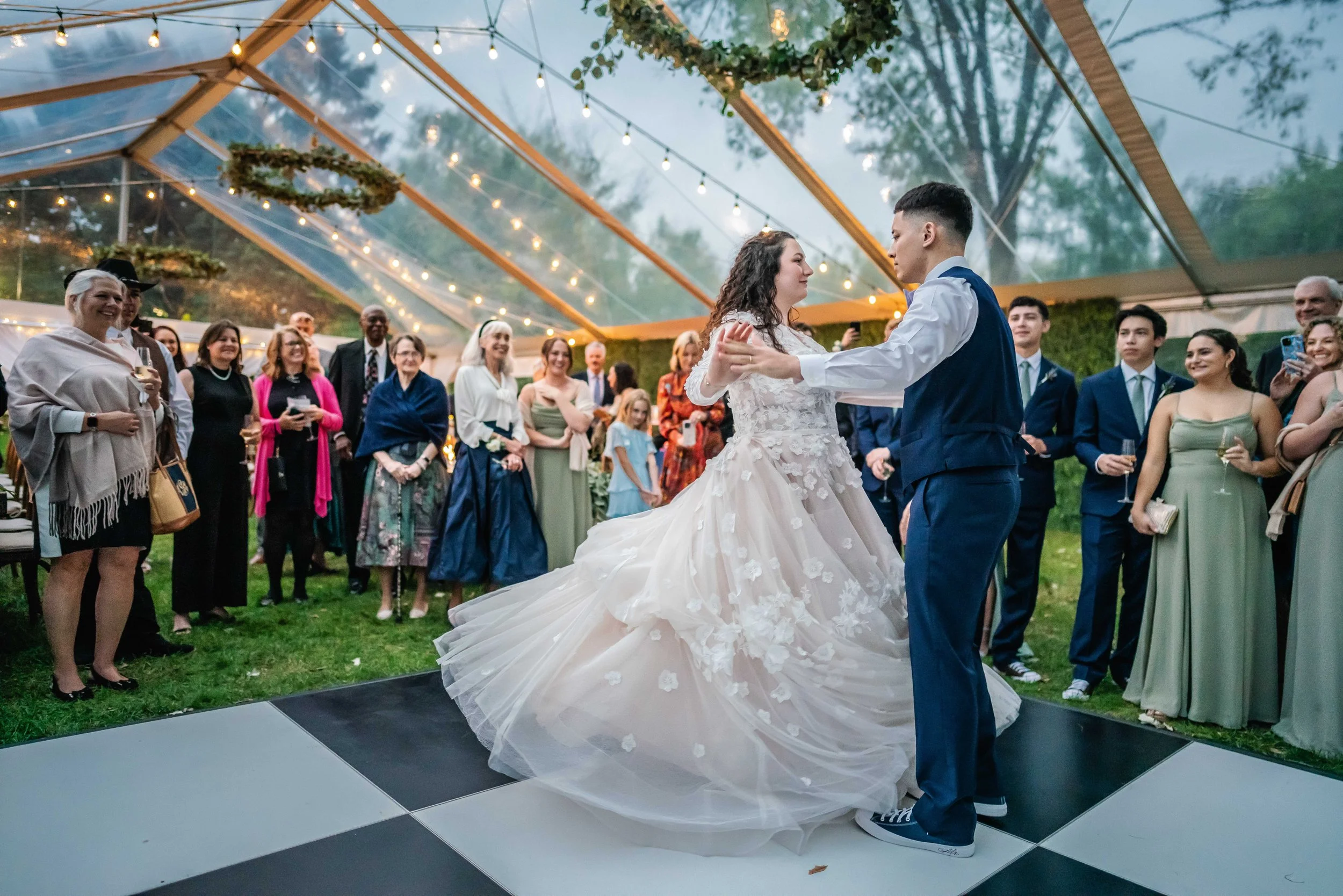Bride and groom dancing at their wedding reception inside a decorated outdoor tent with string lights, surrounded by guests.