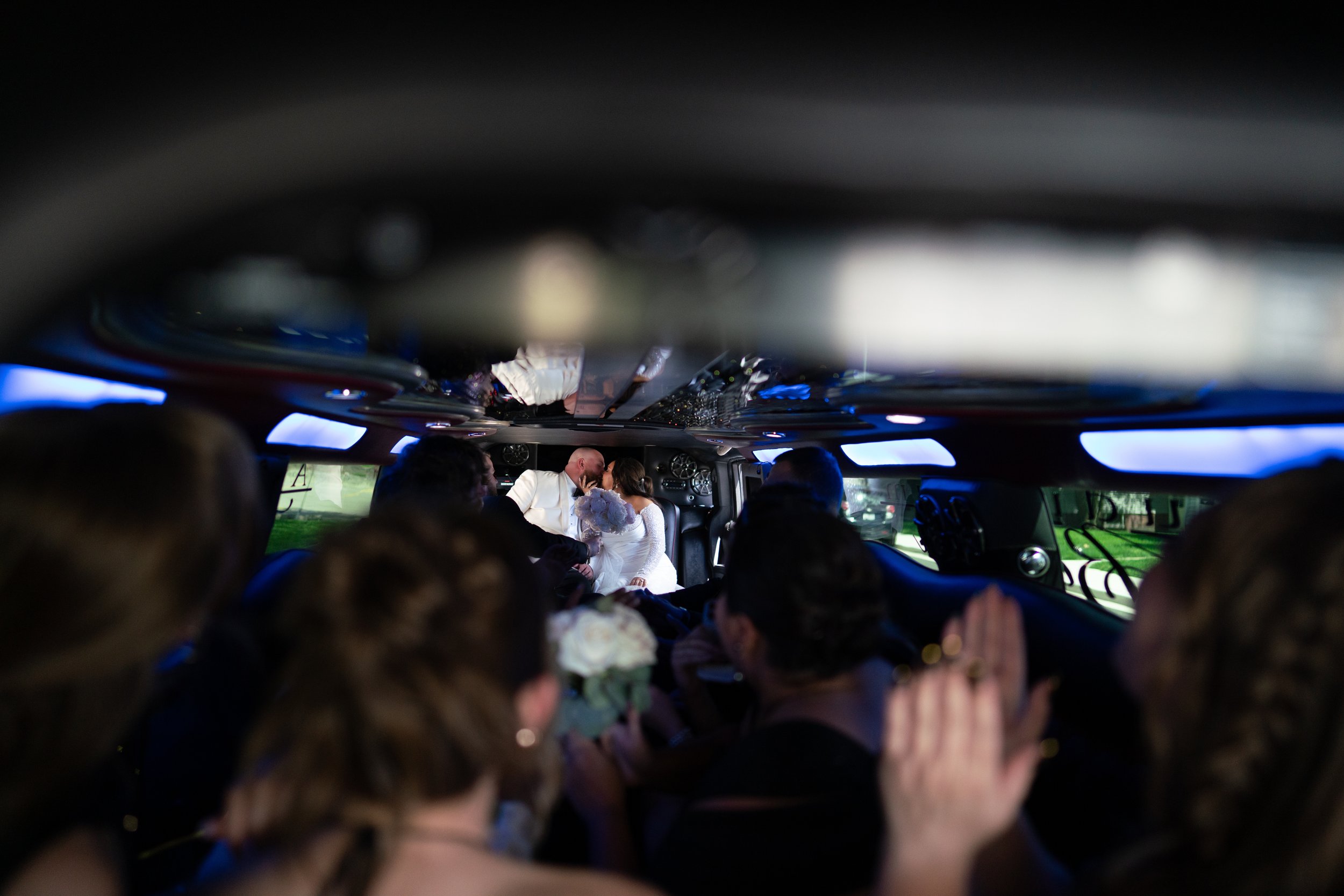 A couple exchanging wedding vows from inside a limousine with friends and family inside, visible through the windows and crowded around.