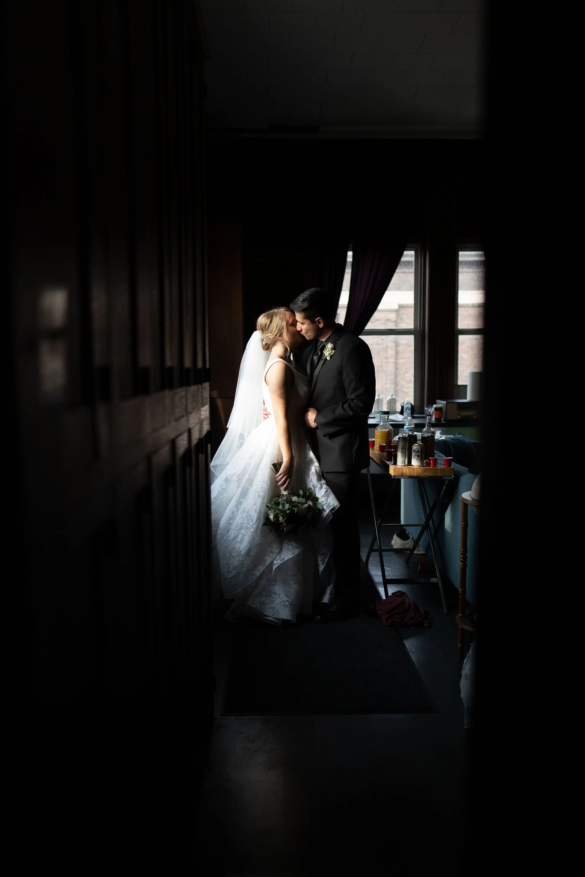 A bride and groom sharing a kiss in a dimly lit room, with sunlight coming through the window behind them, and a bar with various bottles and cups visible on a table next to them.