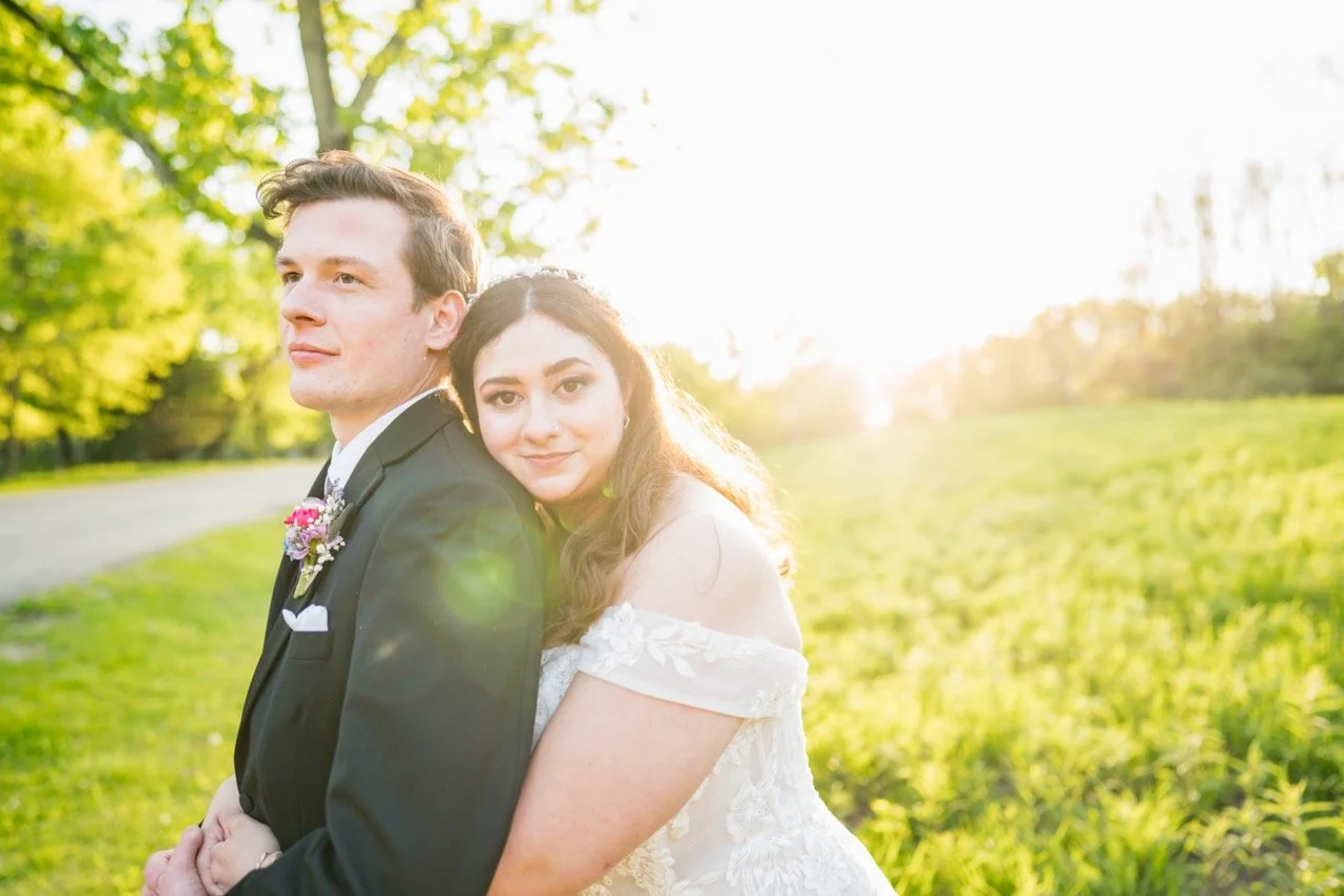 A newlywed couple standing outdoors in a sunny field during golden hour, with the bride leaning on the groom's back, both smiling softly.