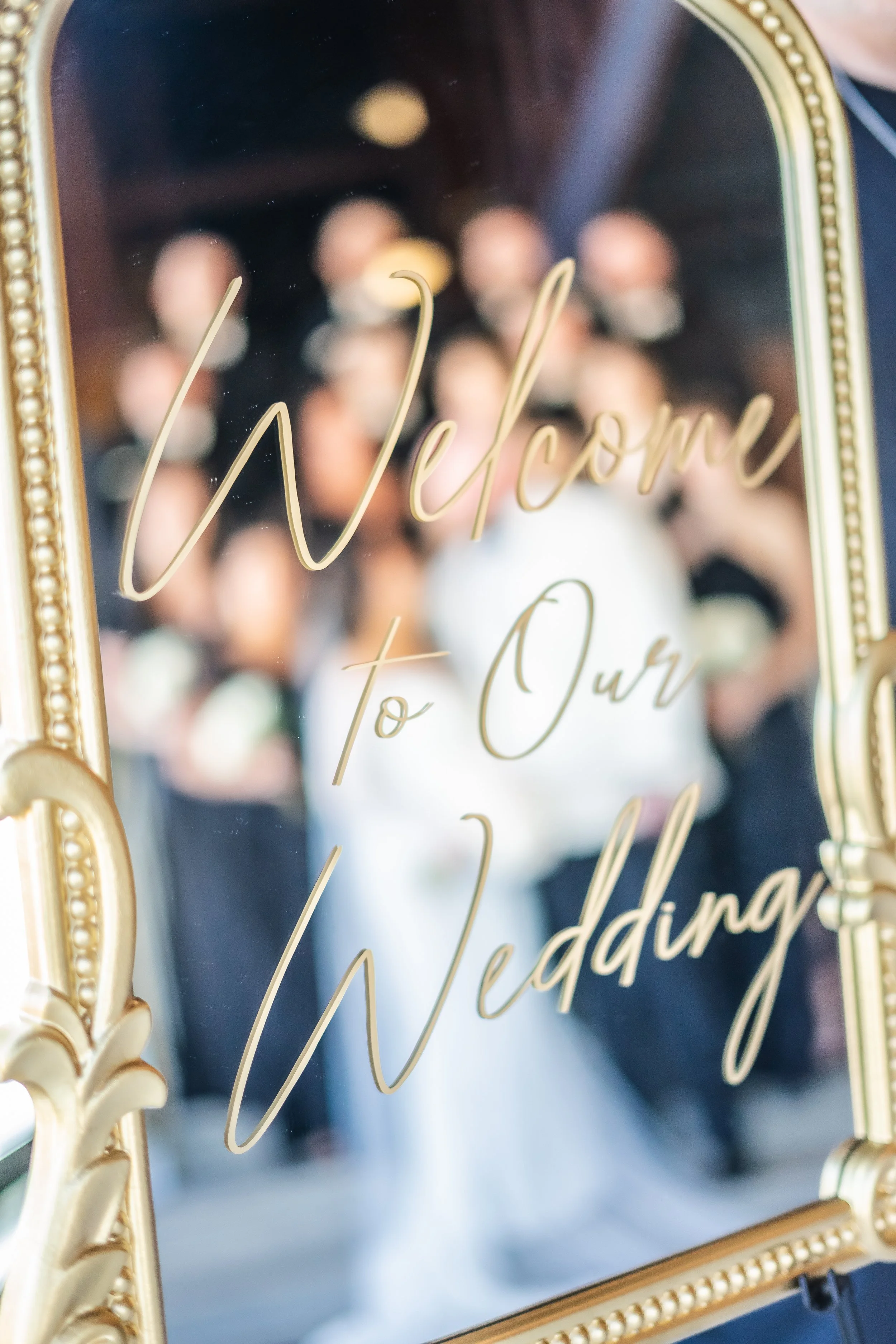 Part of a gold-framed mirror with the word 'Welcome' written in gold cursive on the glass, reflecting blurred faces of people in the background.