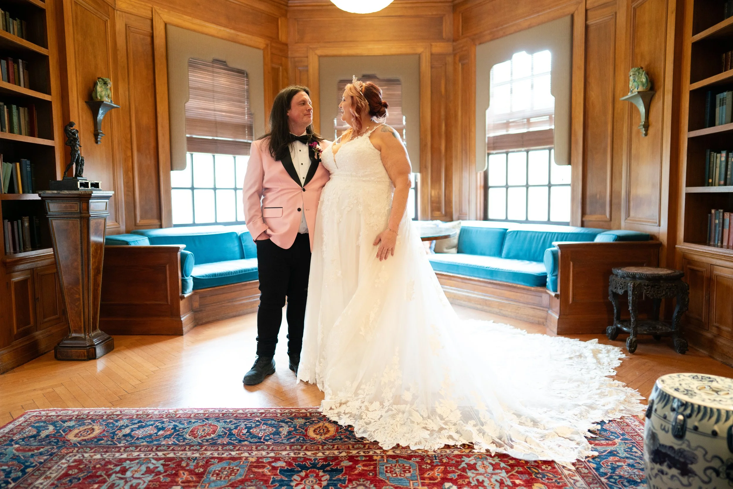 A couple in wedding attire, holding hands and gazing at each other, standing inside a warmly lit room with wood-paneled walls, large windows with blinds, and blue couches in the background.