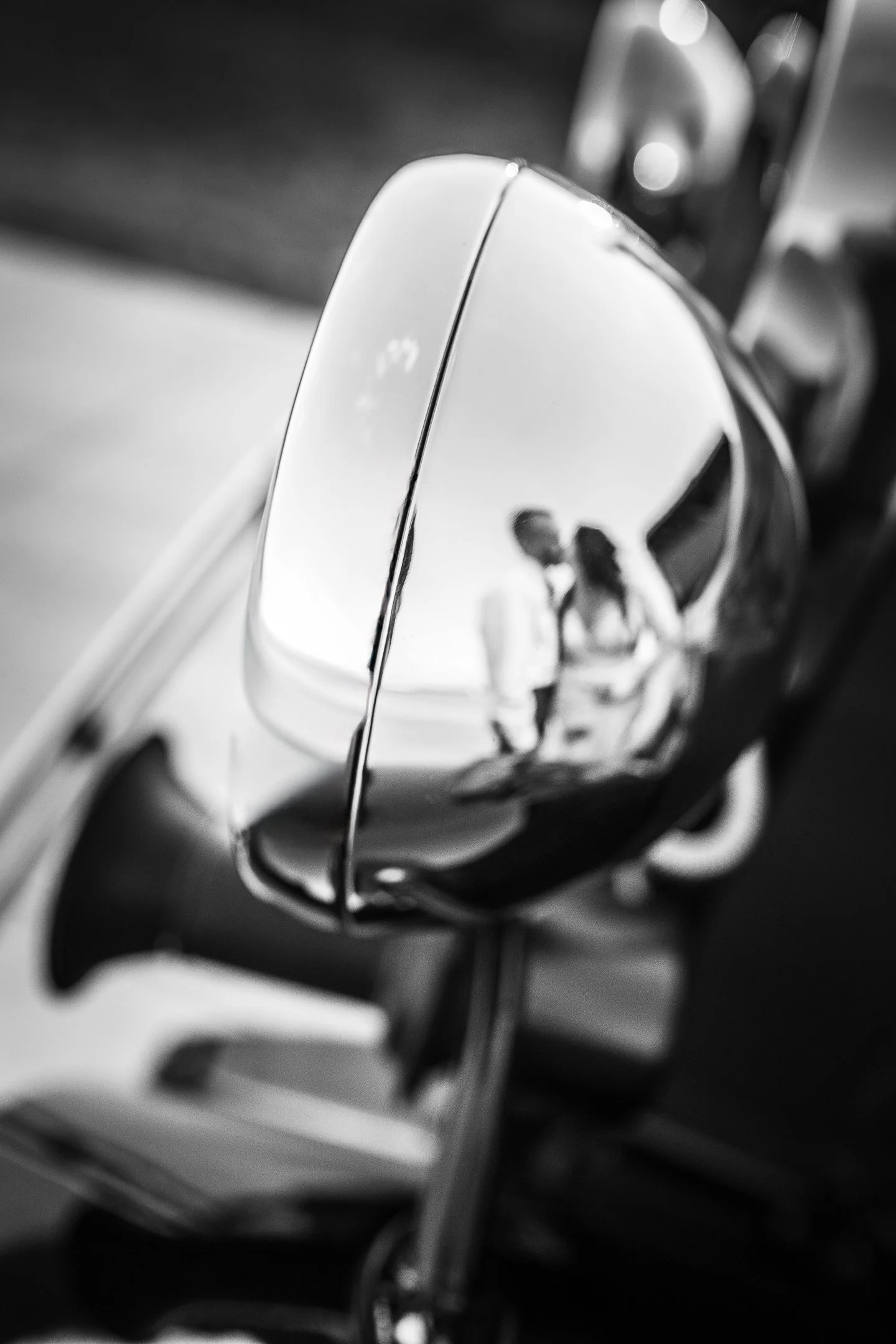 Close-up black and white photograph of a shiny chrome appliance, likely a toaster, reflecting a blurred human figure. The background is out of focus with additional appliances or objects visible.