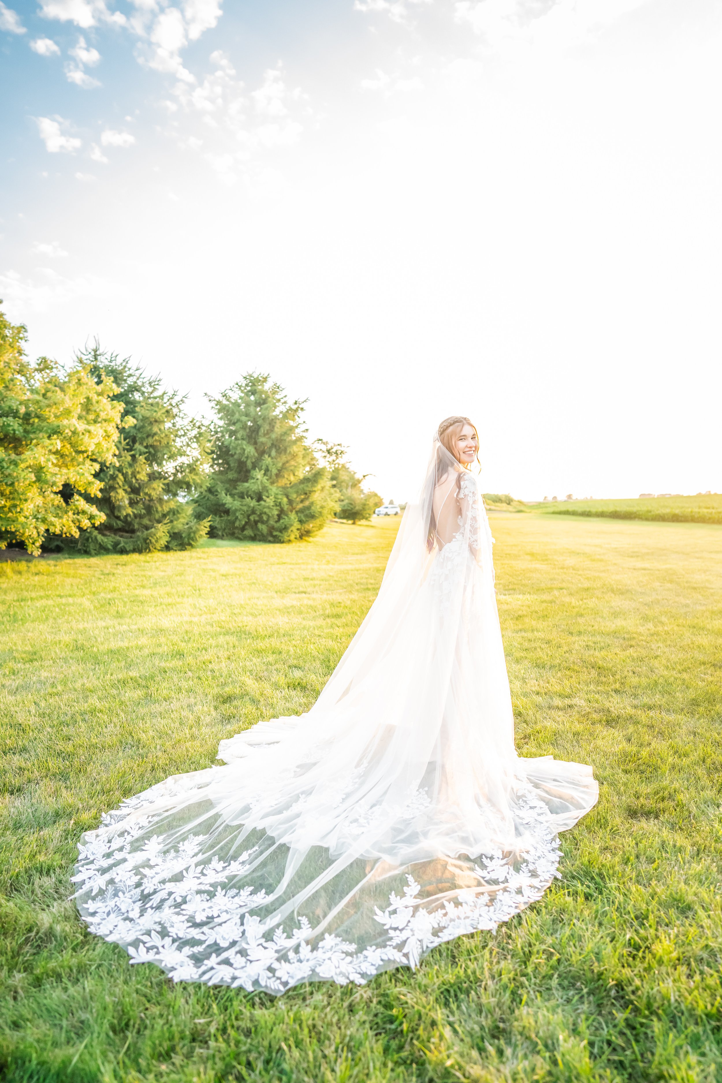 A bride in a white wedding dress with lace details, smiling and standing on a grassy field with trees in the background, under a bright sky.