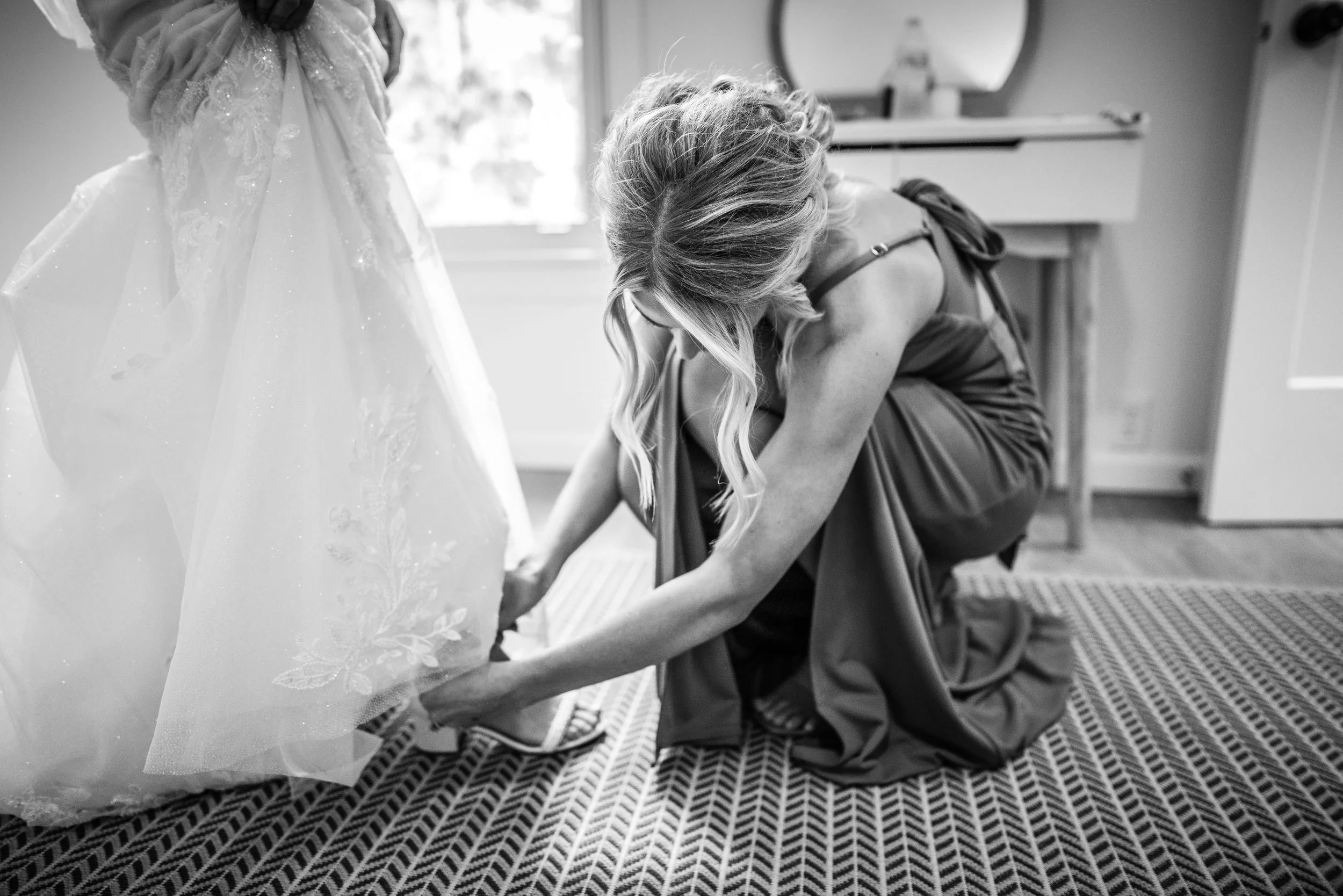 A woman in a formal dress helping a young girl in a gown, both bending down, in a room with natural light, preparing for a special occasion.