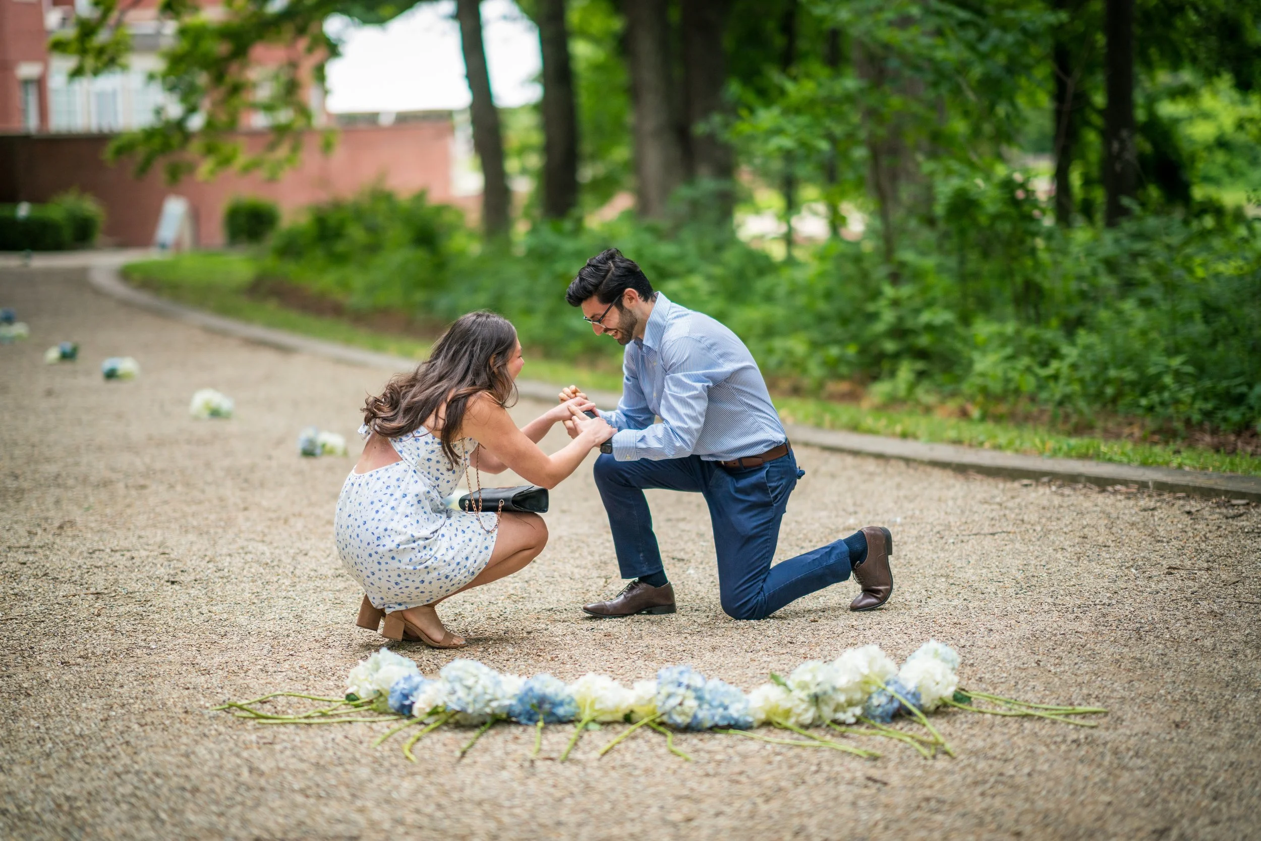 A man kneeling on one knee holding the hand of a woman who is squatting in front of him; they are smiling at each other in an outdoor park setting with trees and flowers around.