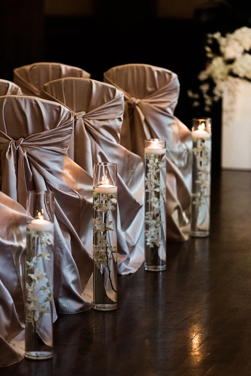 Pink satin chair covers and orchids submerged in cylinder vases lining aisle for wedding ceremony at Minneapolis Club
