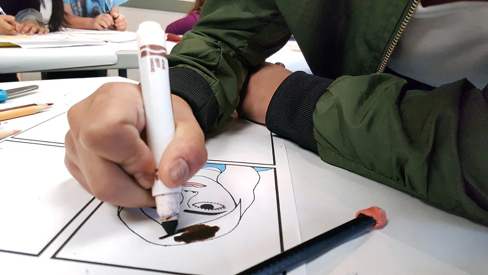 A teen student carefully inks and colors her comic, which is a social commentary on beauty standards.