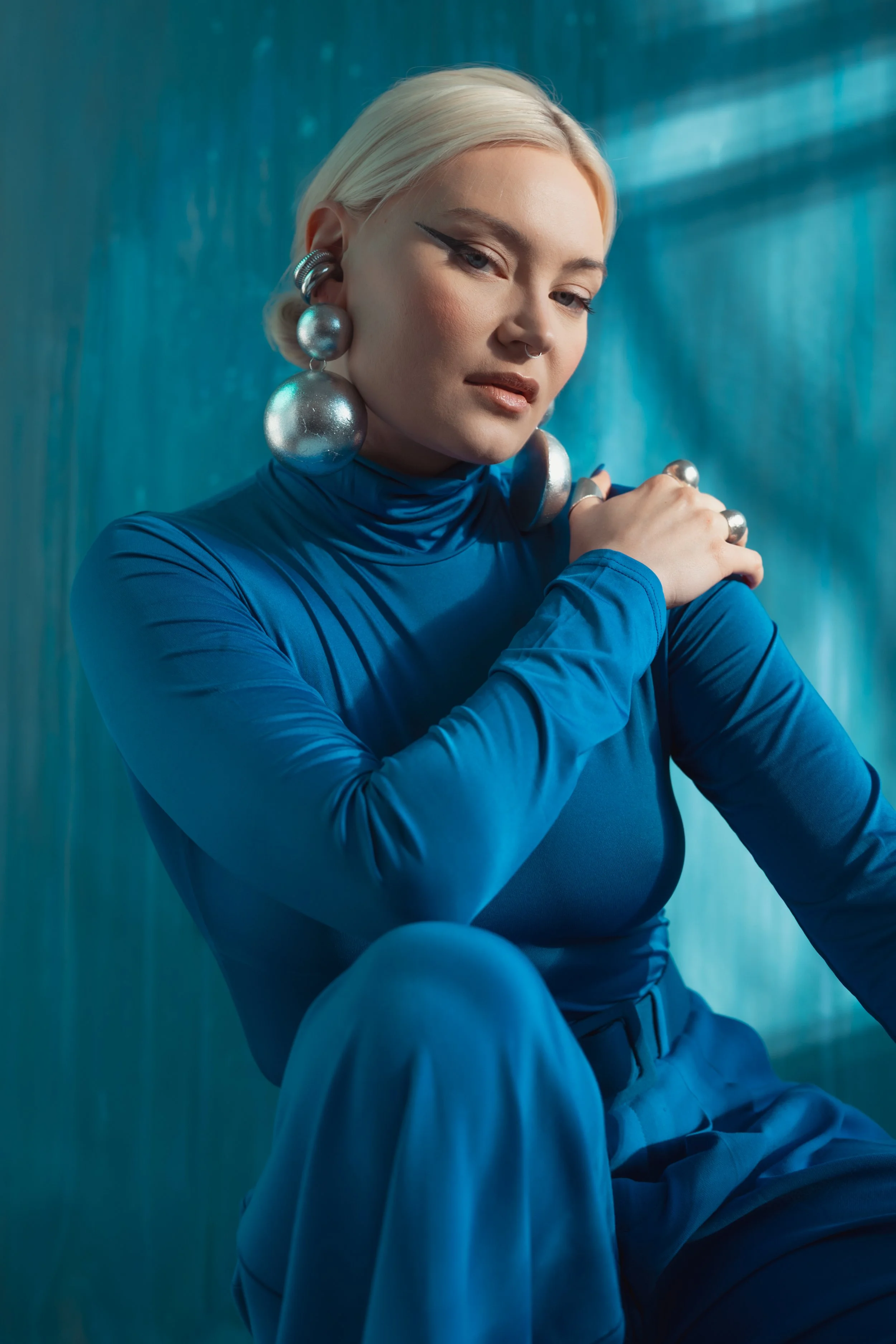 A blonde woman dressed in a blue high-neck top and pants, wearing large silver earrings and rings, sitting against a blue textured background with a contemplative expression.