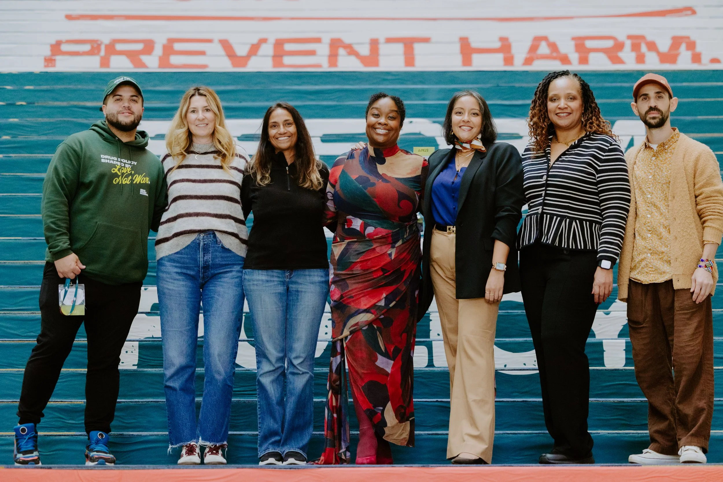 Group of eight diverse people standing on stairs in front of a mural with the words 'PREVENT HARM' visible at the top.
