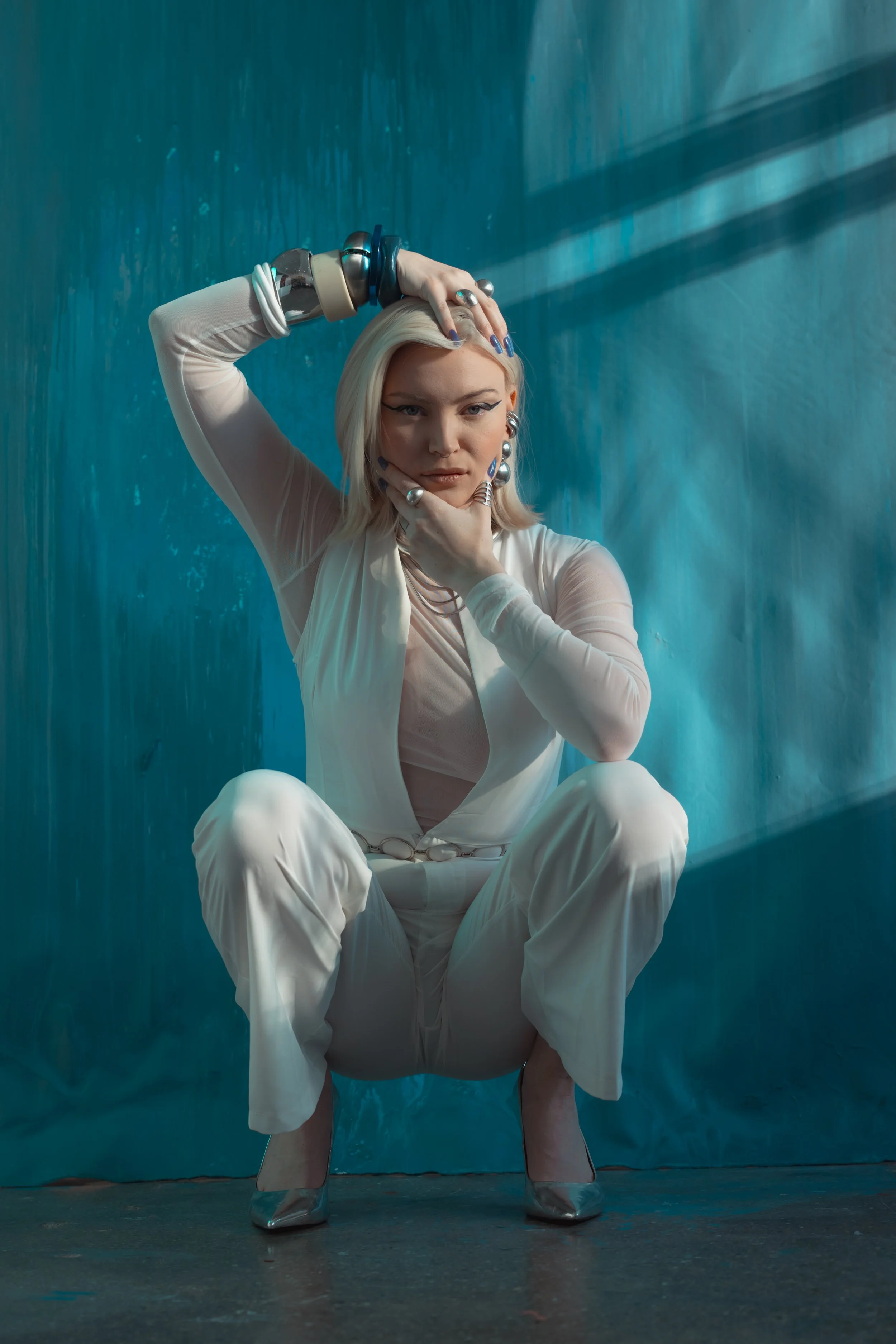 A woman with platinum blonde hair dressed in white, wearing jewelry, squatting against a teal backdrop with light and shadow patterns.