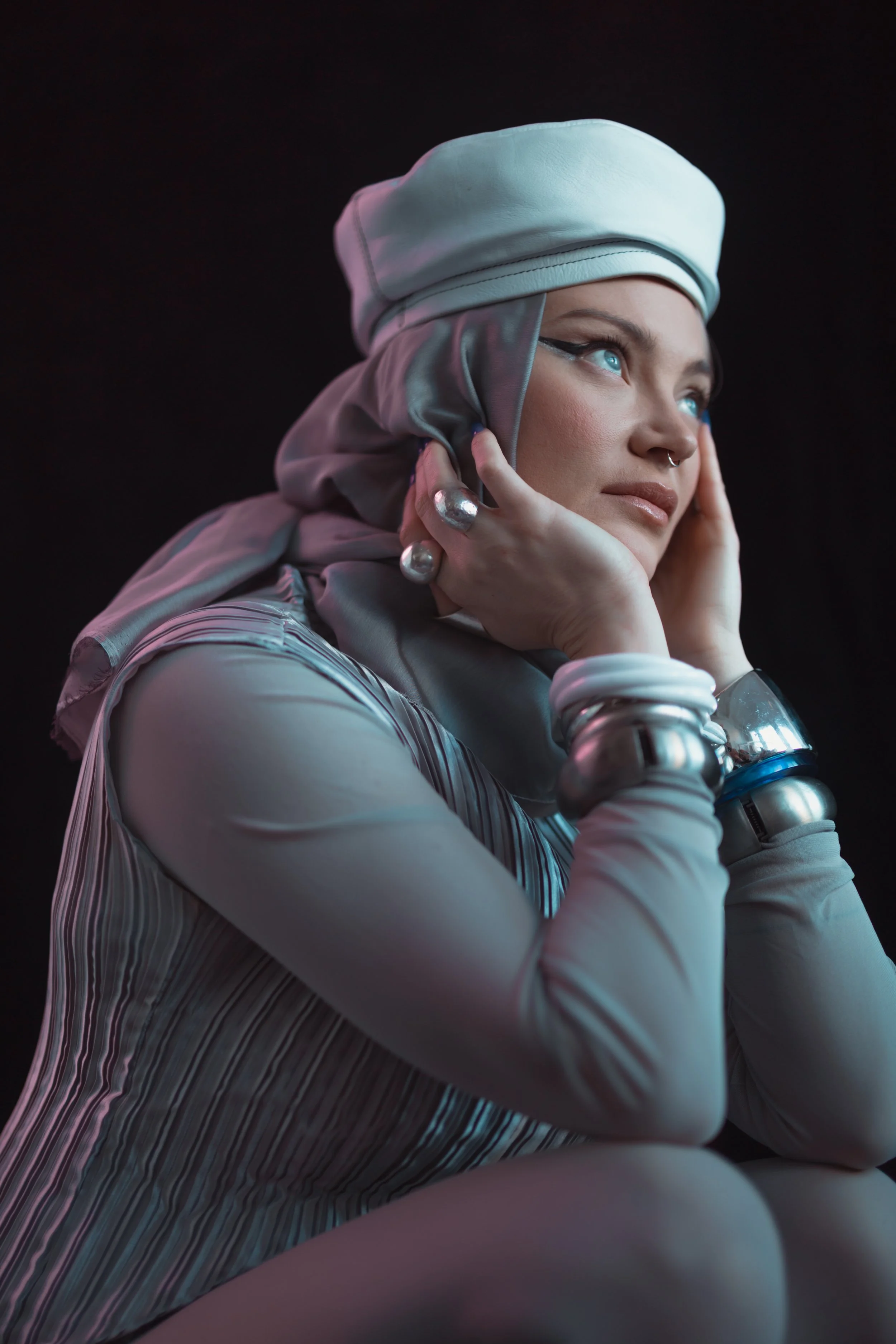 A woman with blue eyes, platinum blonde hair, and metallic jewelry rests her face in her hands, wearing a white head covering and a striped, long-sleeve top, against a dark background.