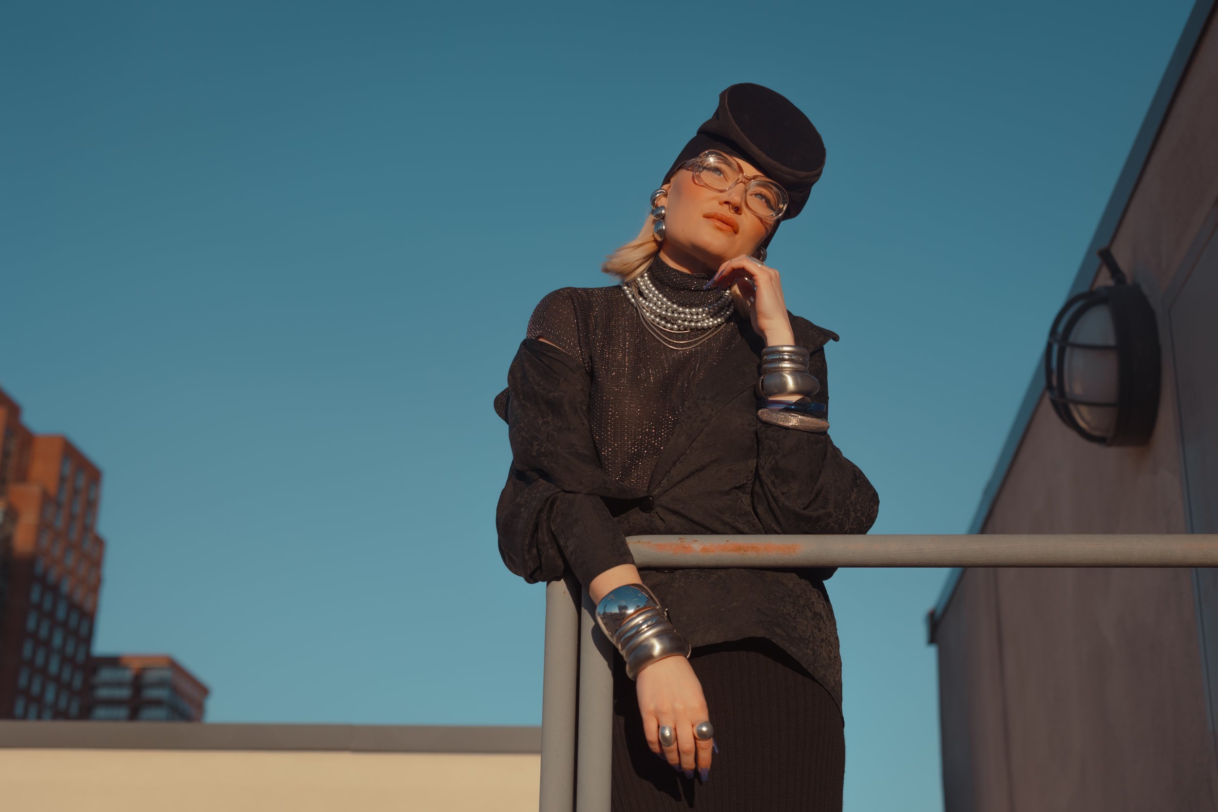 A stylish woman on a rooftop wearing a black outfit, multiple silver jewelry pieces, a black hat, and glasses, standing against a blue sky during sunset.