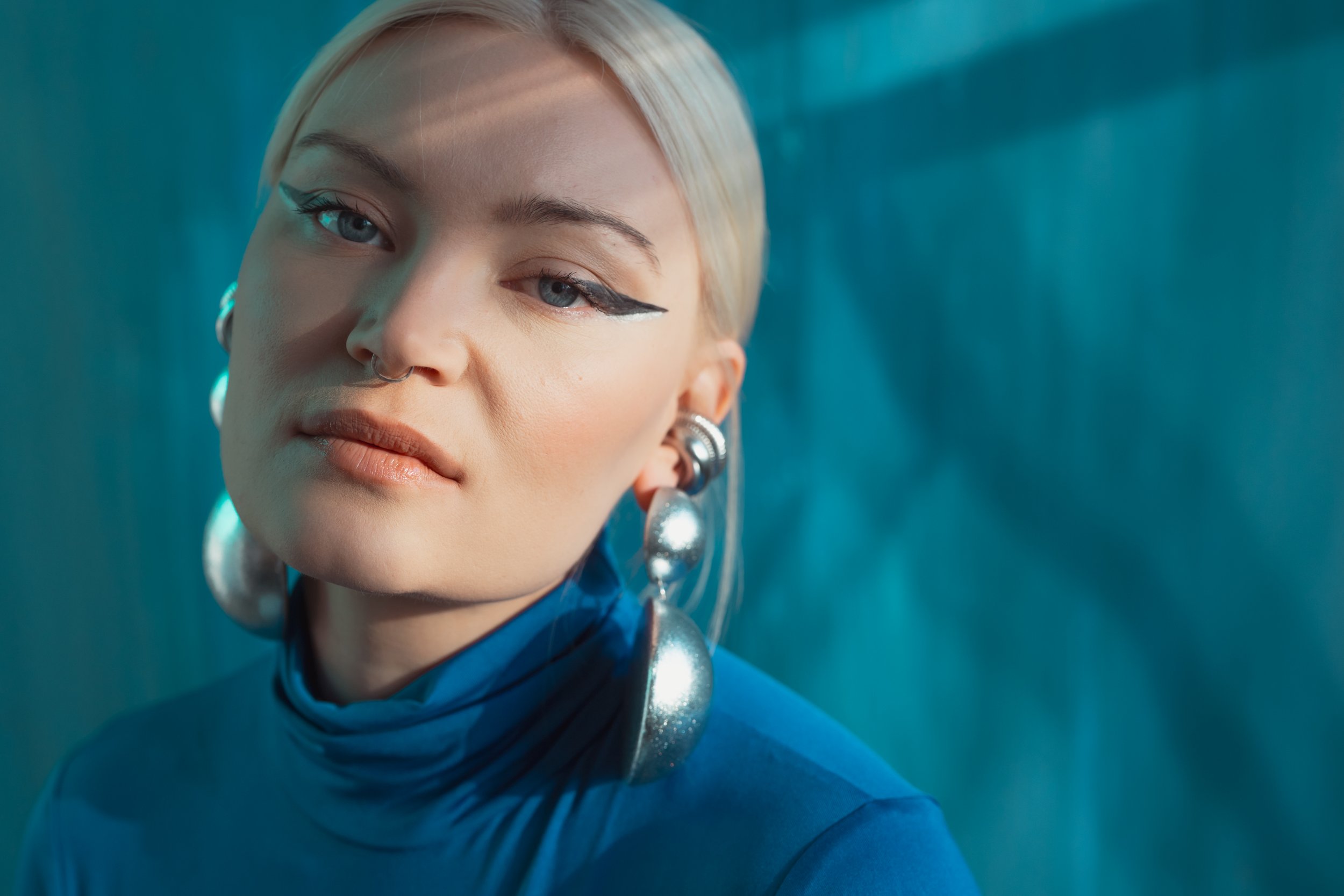 Close-up of a young woman with platinum blonde hair, blue eyes, bold eyeliner, and silver earrings, posing against a textured teal background.