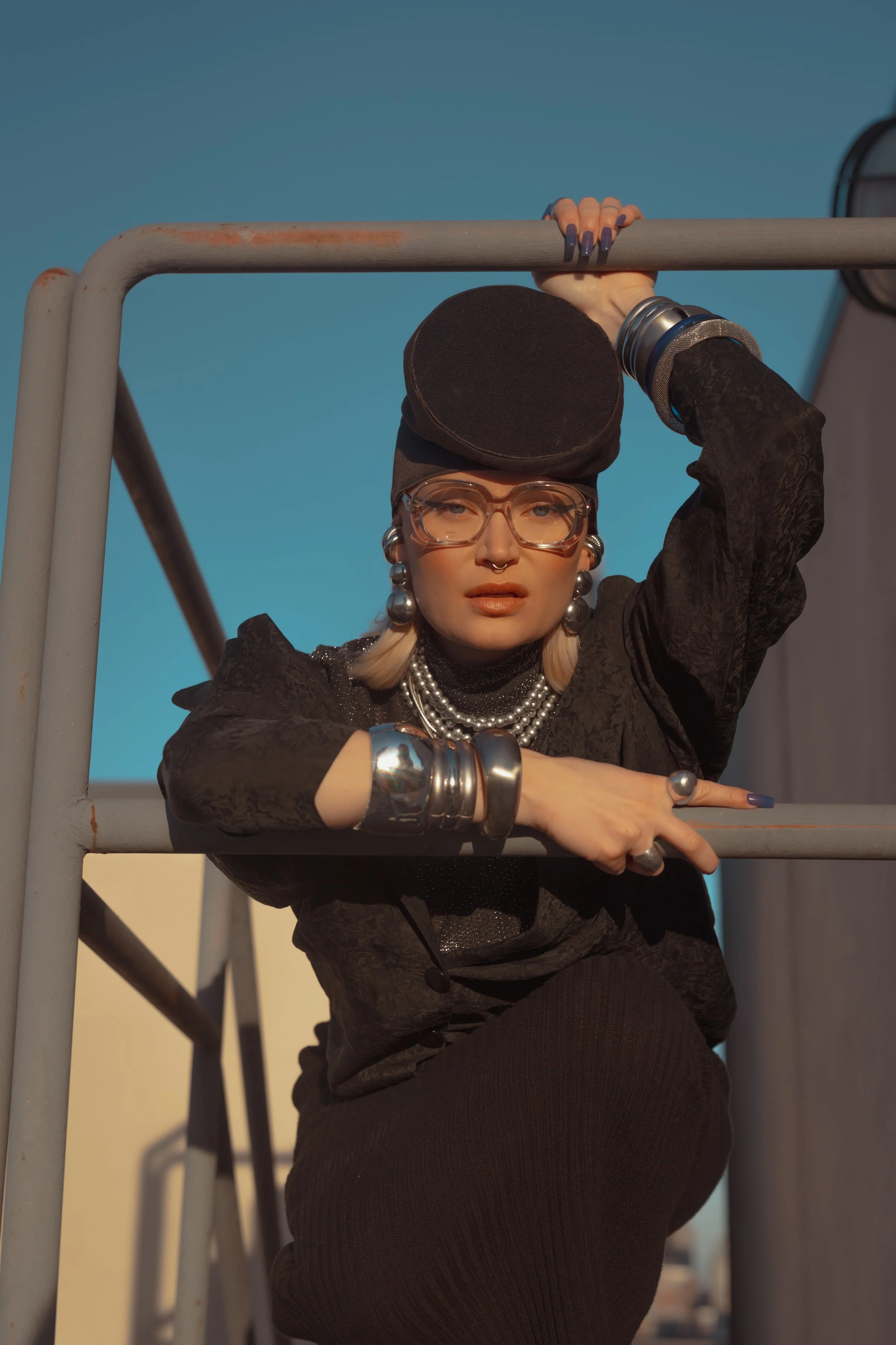 A woman with blonde hair and tattoos on her hand posing on a rooftop with a clear blue sky background, wearing large glasses, jewelry, and fashionable clothing.