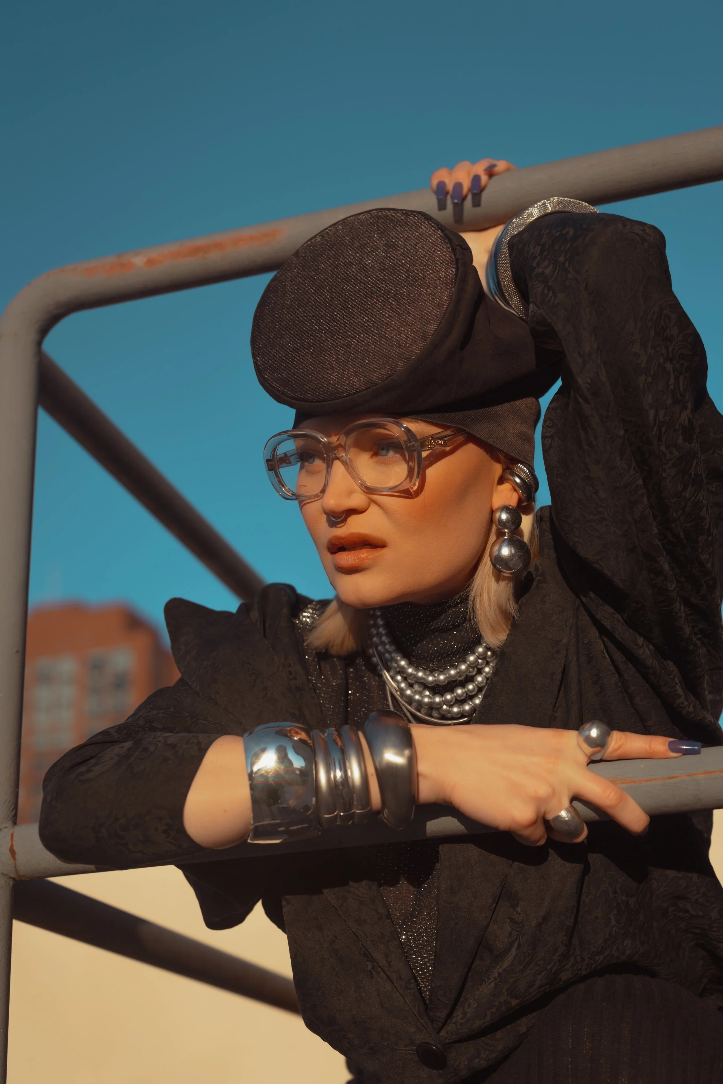 A woman wearing large glasses, pearl jewelry, and silver accessories gazes into the distance while leaning on a metal railing.