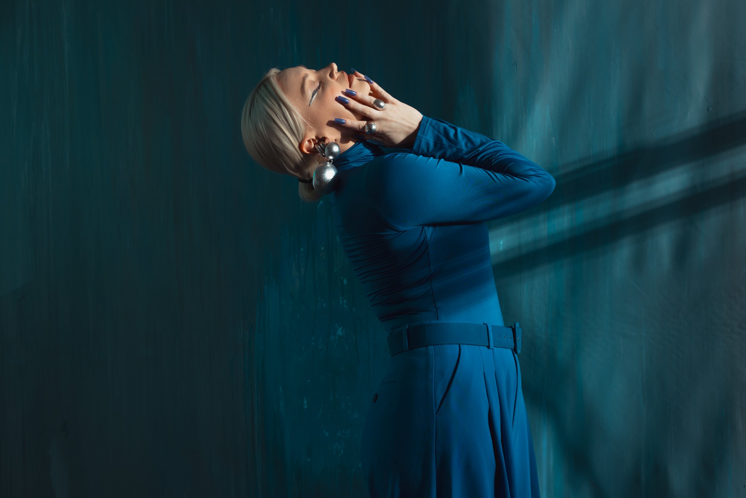 A woman with blond hair styled in a bun wearing a blue dress, large silver earrings, and multiple rings, standing against a dark background with moody lighting.