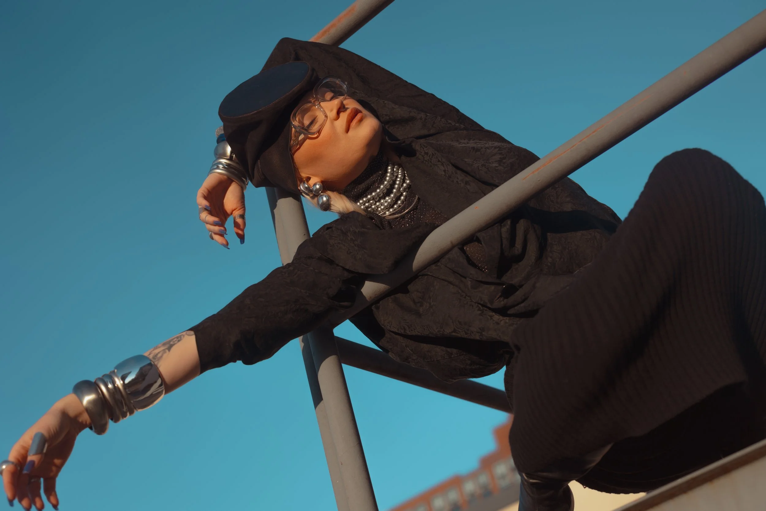 A woman with platinum blonde hair, wearing black glasses, jewelry, and all black clothing, is leaning back and resting on a ladder against a clear blue sky. She appears relaxed and fashionable.