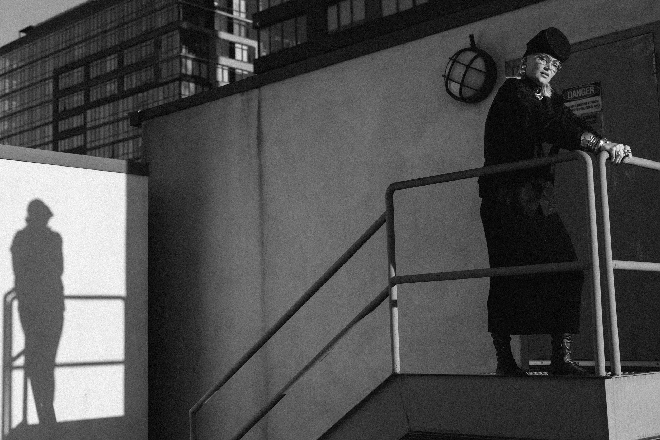 A woman standing on a balcony in an urban setting, with her shadow cast onto a blank wall to her left.