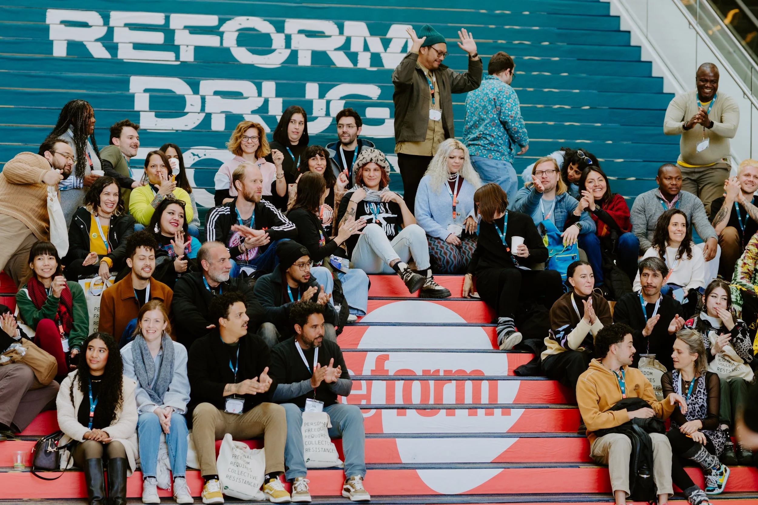 A diverse group of people sitting on red and blue steps at a conference or event, with some clapping, smiling, and talking. The background features large text that reads 'REFORM' and 'DRUG'.