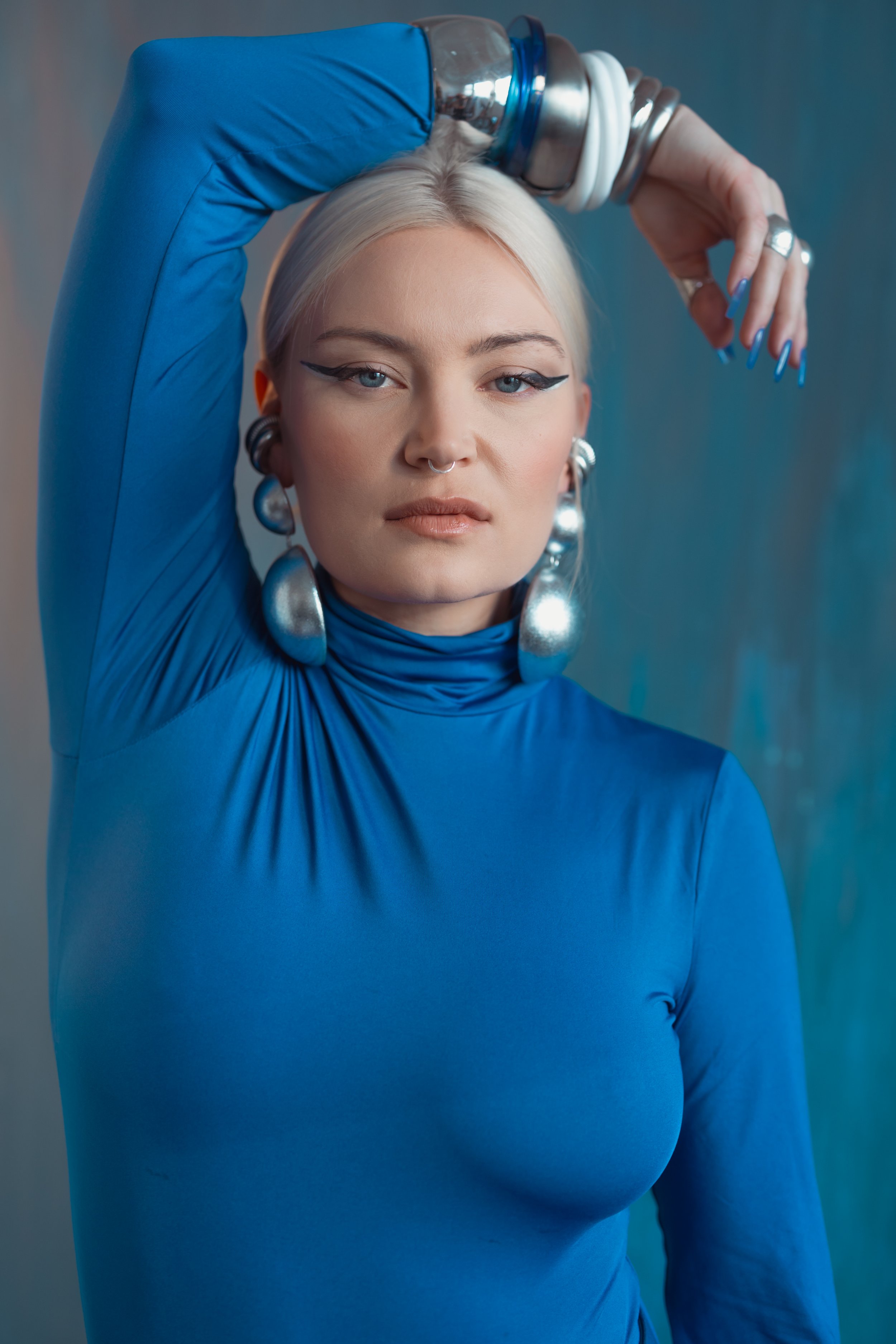 A woman with platinum blonde hair and bold makeup, wearing large silver earrings and a matching septum ring, dressed in a form-fitting blue turtleneck, posing with her arm raised and supporting a metallic and blue water bottle on her head.
