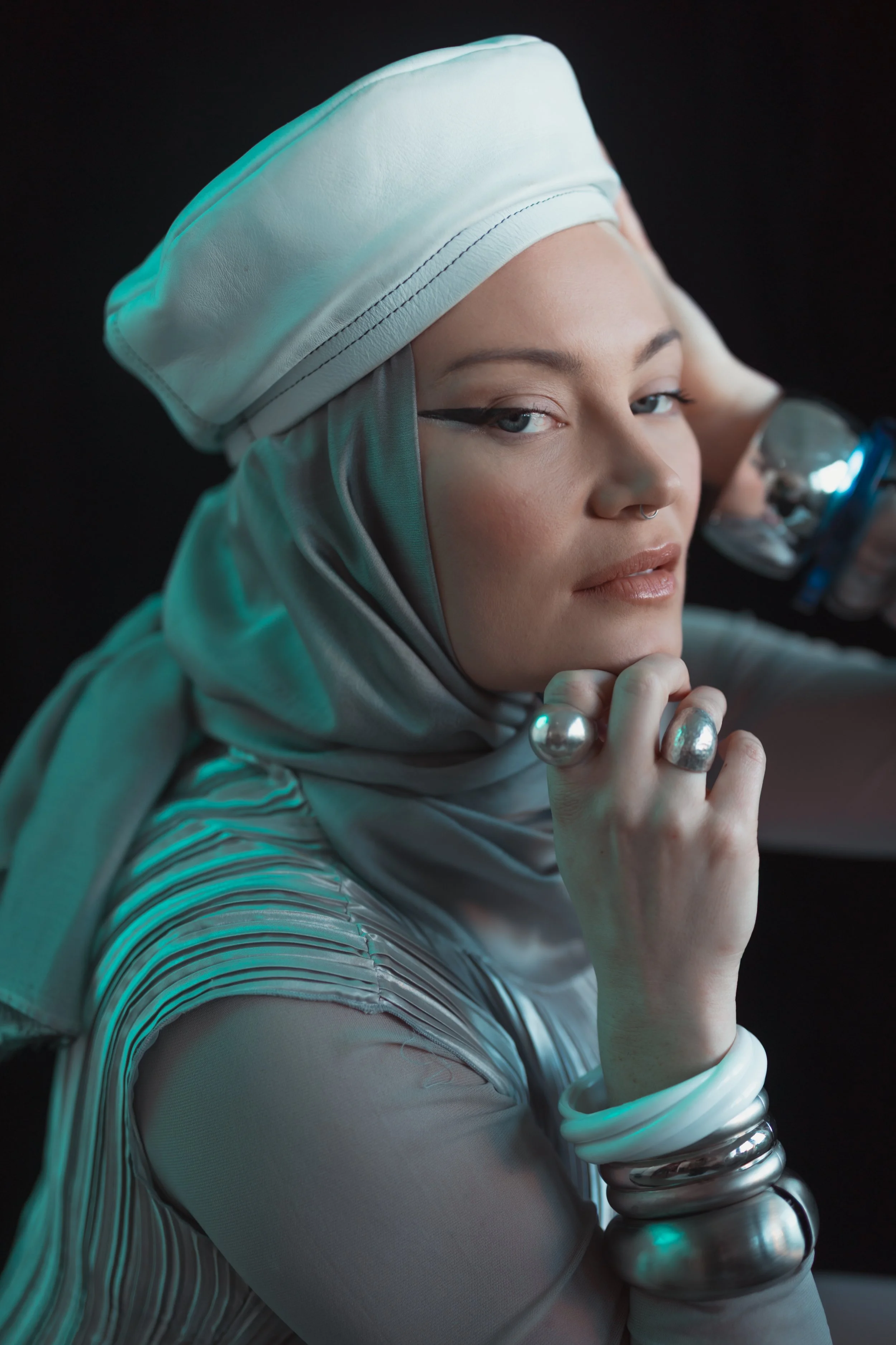 A woman with light makeup, silver jewelry, and a headscarf, dressed in futuristic clothing, holding a metallic sphere and a bottle, with her head tilted and looking at the camera against a dark background.