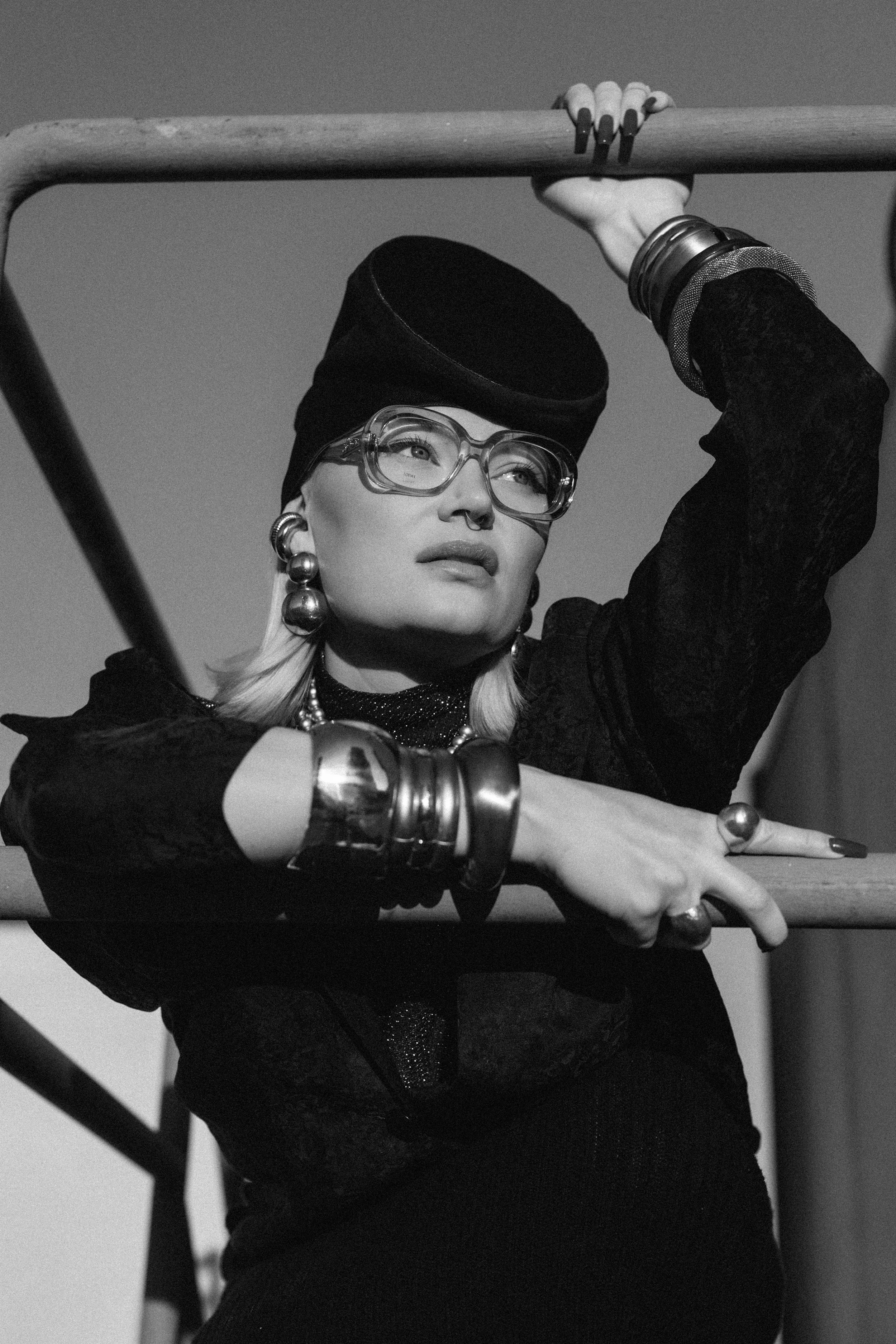 Black and white portrait of a woman with blonde hair styled back, wearing oversized glasses, large metallic jewelry, and a dark head wrap, posing with her arm resting on a railing.