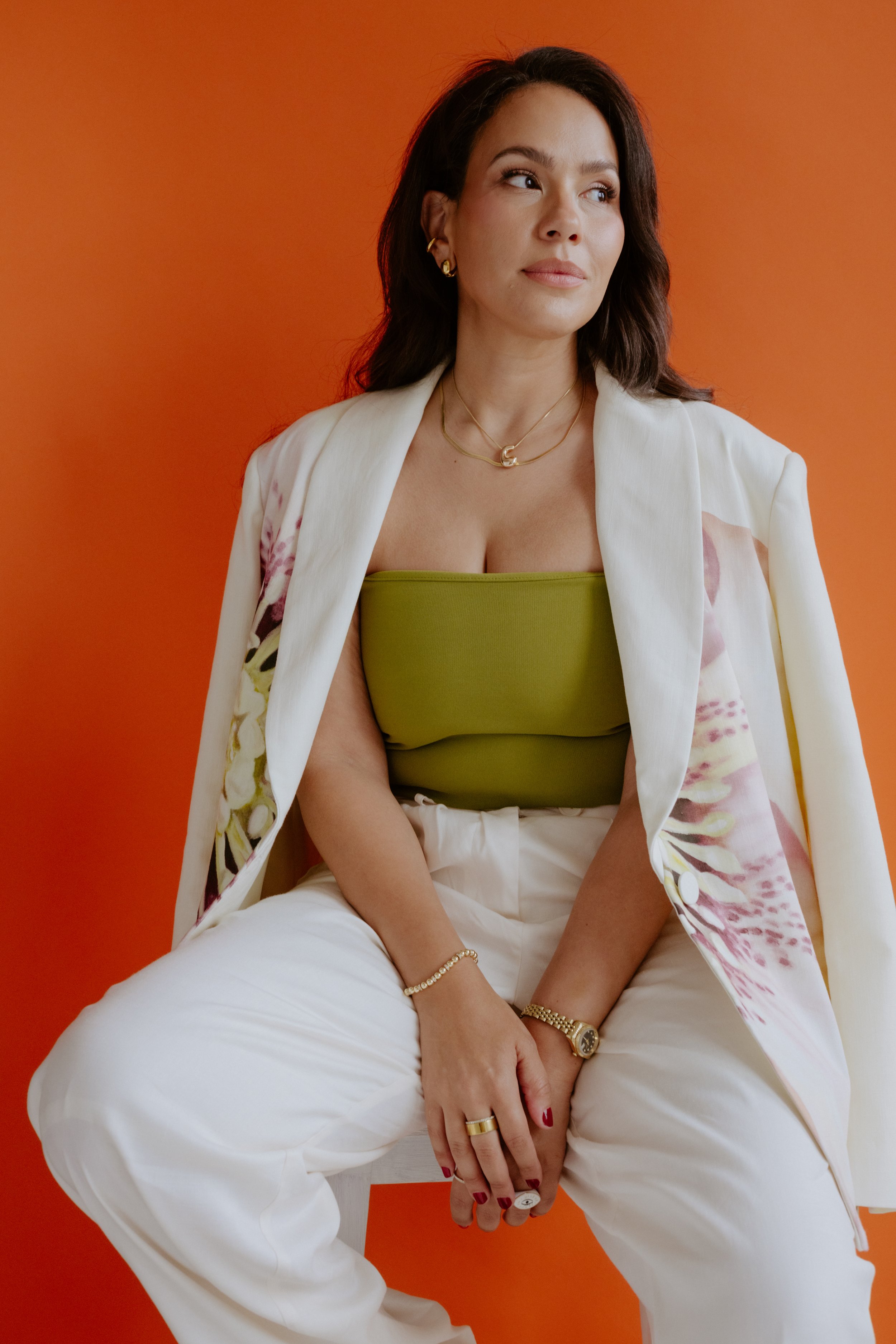 A woman with dark hair wearing a green strapless top and white pants, seated against an orange background. She has a floral blazer draped over her shoulders, gold jewelry, and red nail polish.