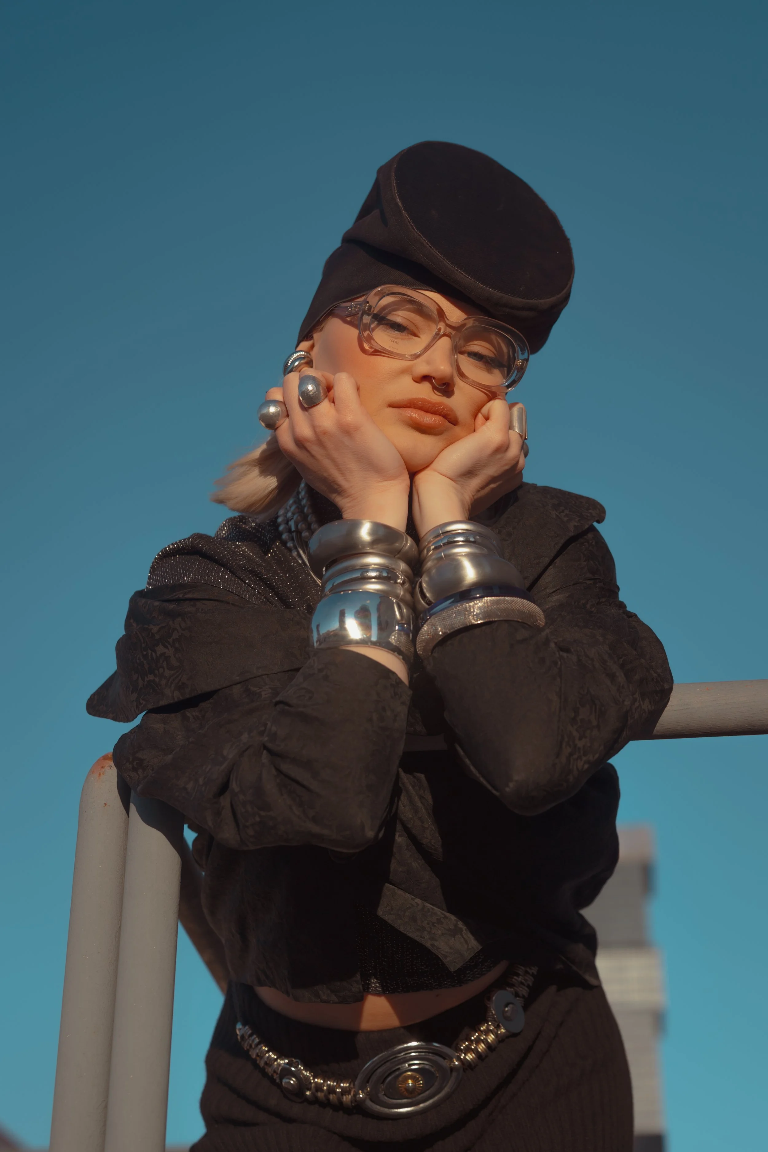 A woman with glasses and multiple silver jewelry, including rings and bracelets, leans on her hands with a thoughtful expression. She is wearing a black hat and a black jacket, and the background shows a clear blue sky.