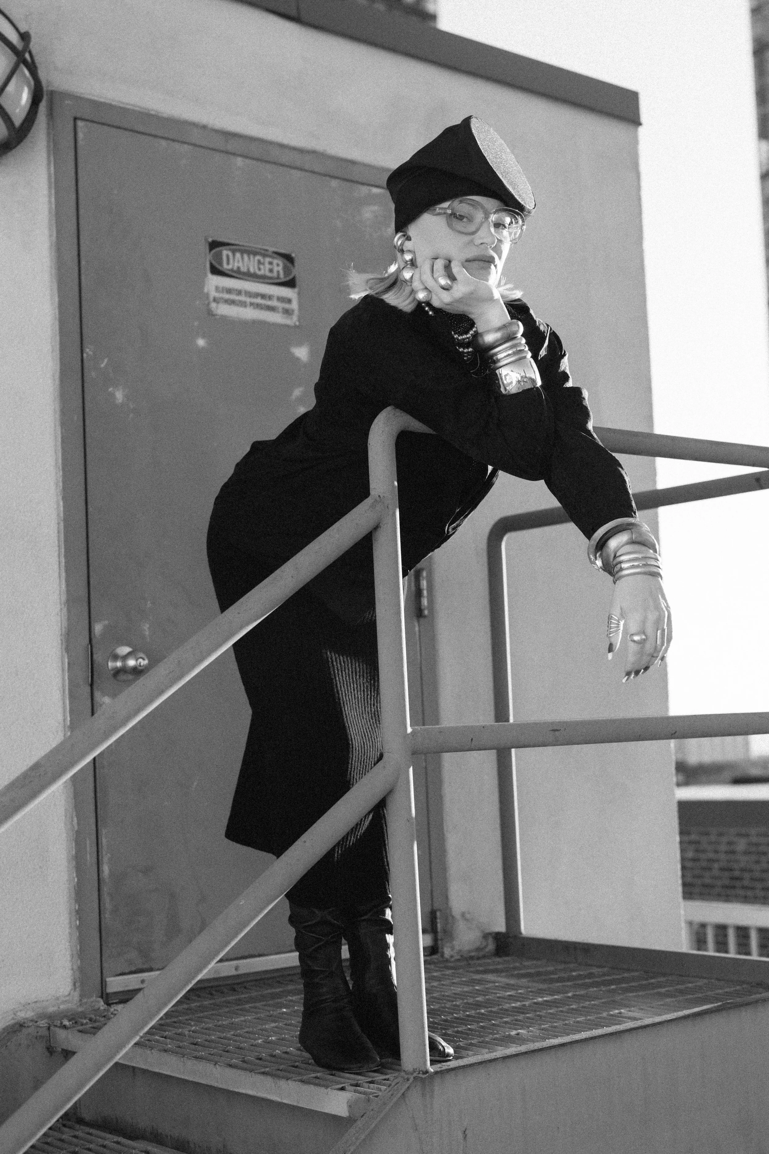 A woman with blonde hair, wearing glasses, a beret, and jewelry, leaning on a metal railing on a rooftop or fire escape, with a closed door and a "DANGER" warning sign in the background.