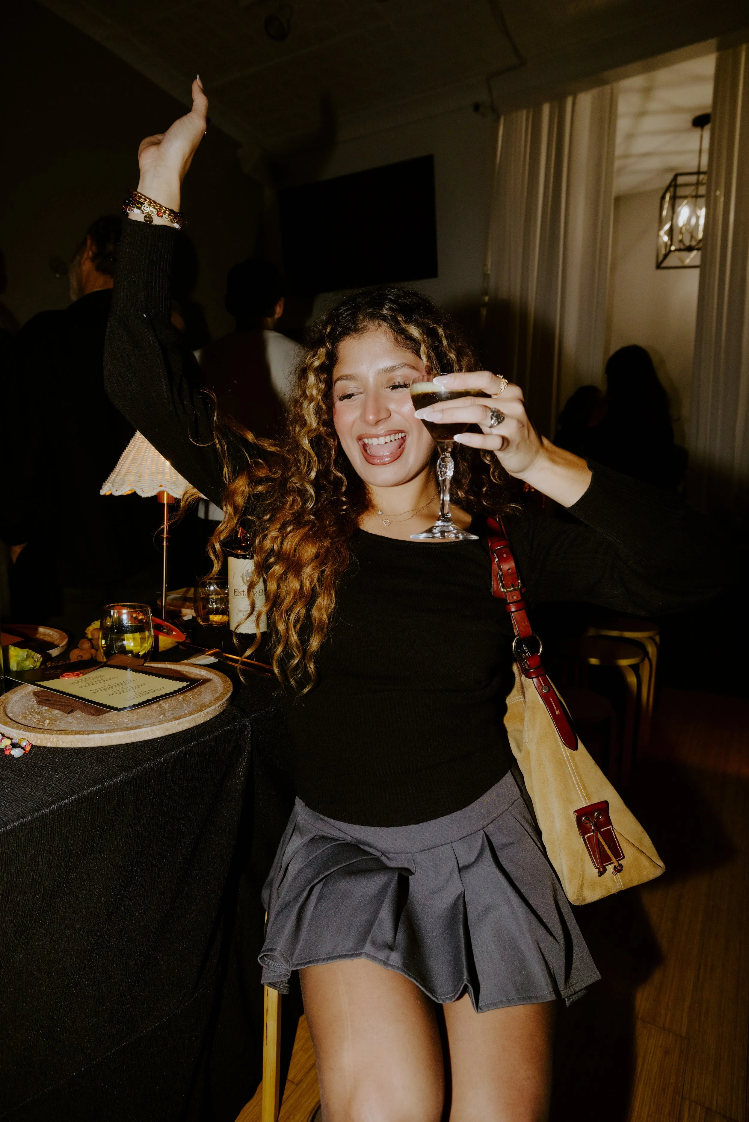 A young woman with curly hair enjoying a drink at a lively party or bar, dancing with her eyes closed and tongue out, holding a glass of dark liquid, wearing a black top and a short gray skirt, with a beige purse over her shoulder. There are other pe