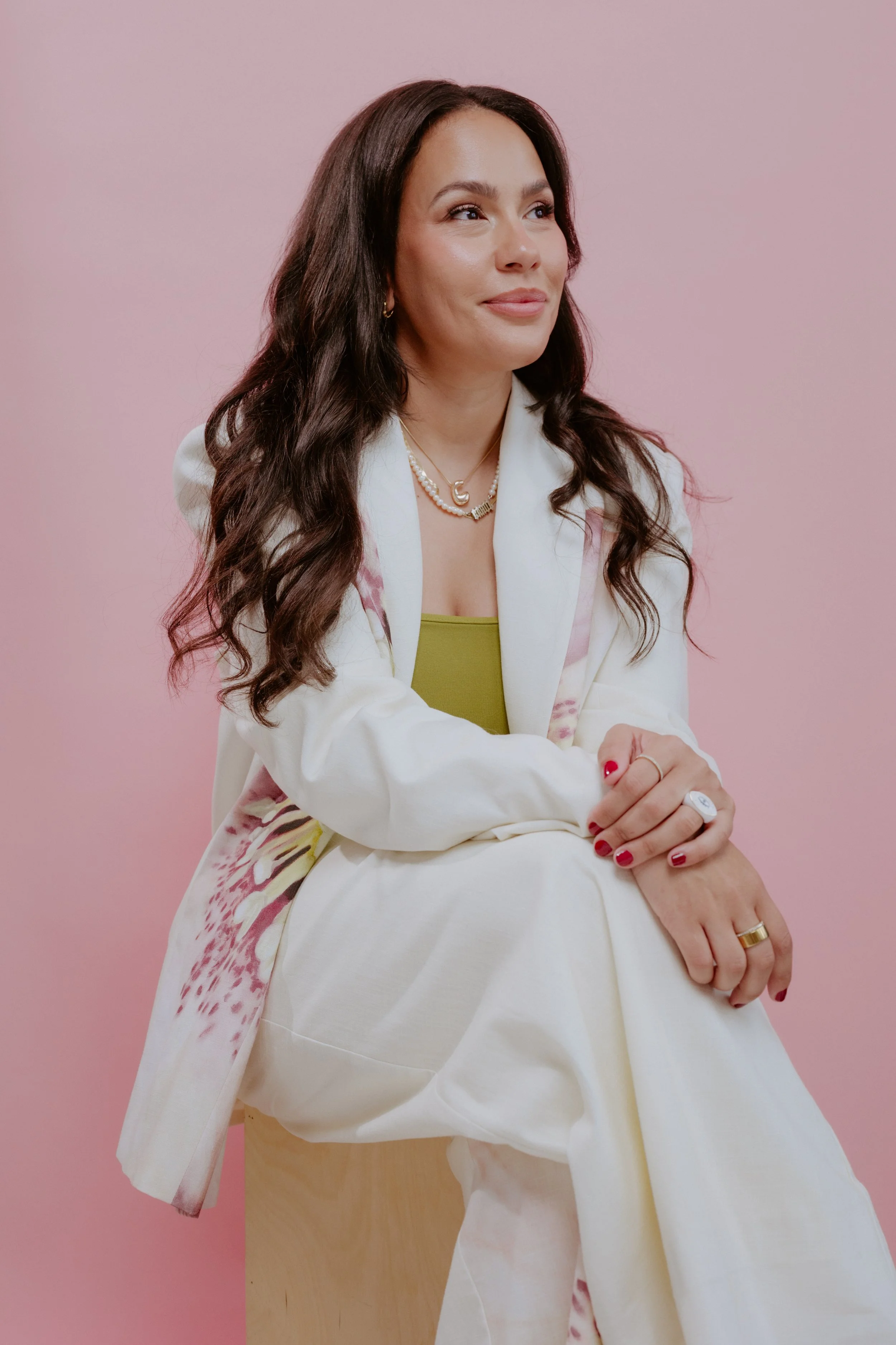A woman with long wavy brown hair wearing a white suit with pink and yellow accents, sitting on a wooden stool against a pink background.
