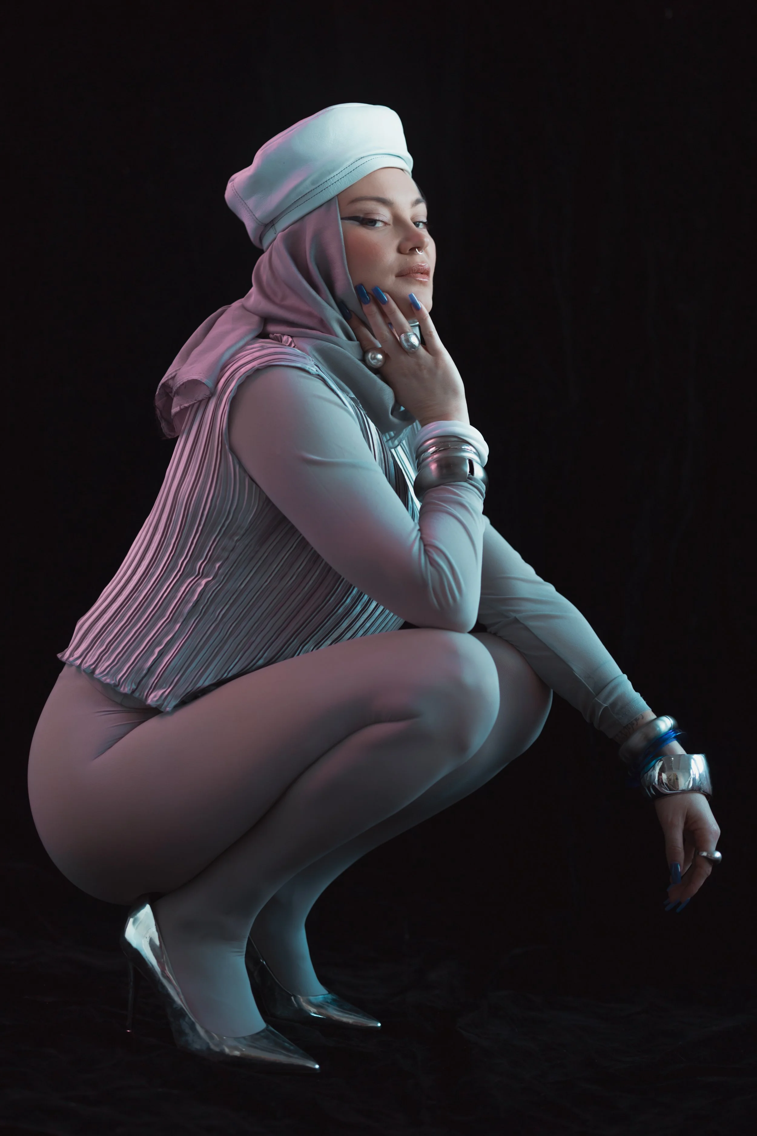 A woman squatting in a fashion shoot, dressed in a headscarf, striped top, and metallic high heels, with jewelry and a dark background.