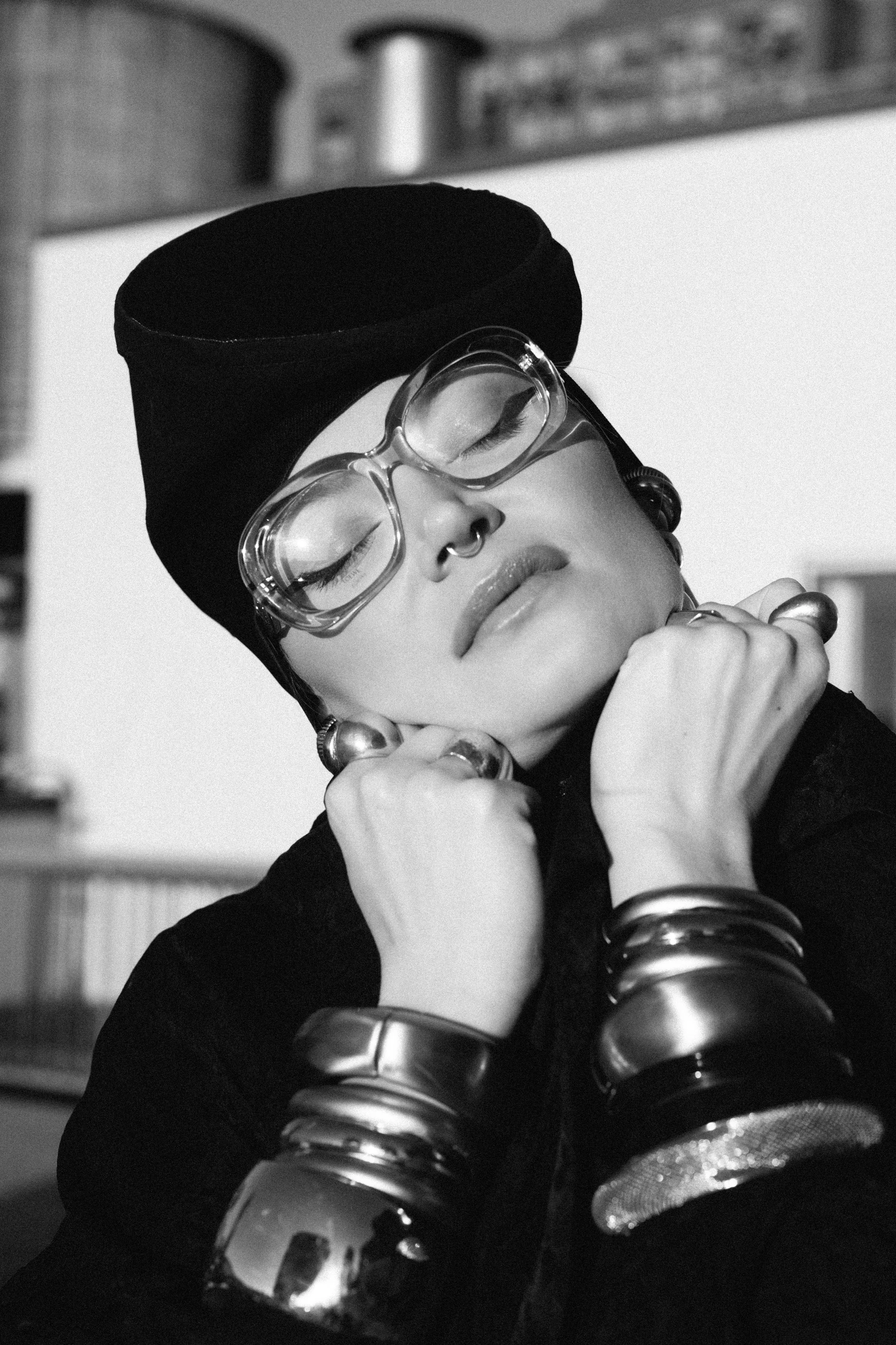 A black and white photo of a woman with her eyes closed, wearing glasses, a head wrap, and multiple bracelets, resting her face on her hands.