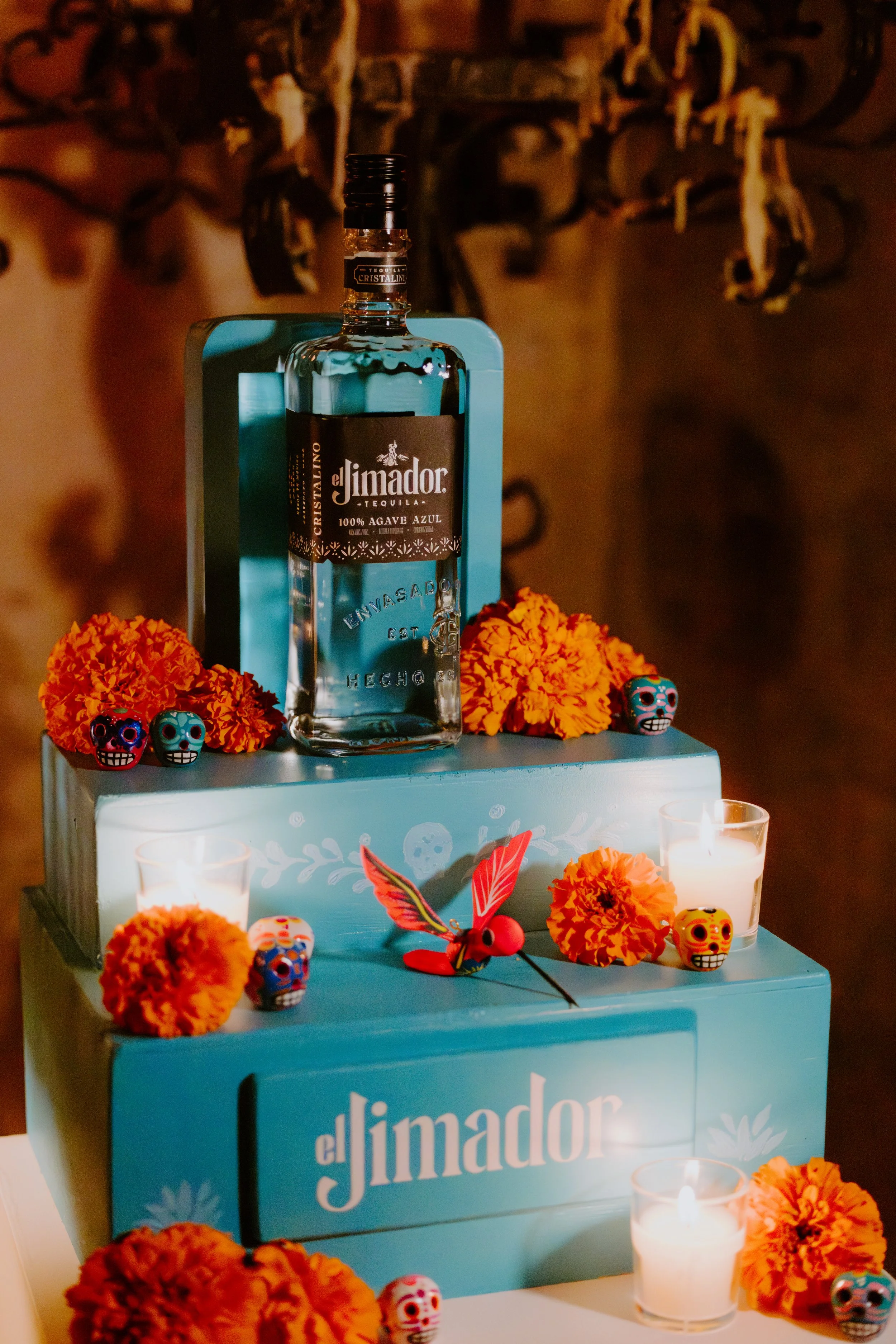 A decorative display featuring a turquoise-themed cake with orange marigold flowers, colorful sugar skull figurines, small candles, and a bottle of El Jimador tequila on top.