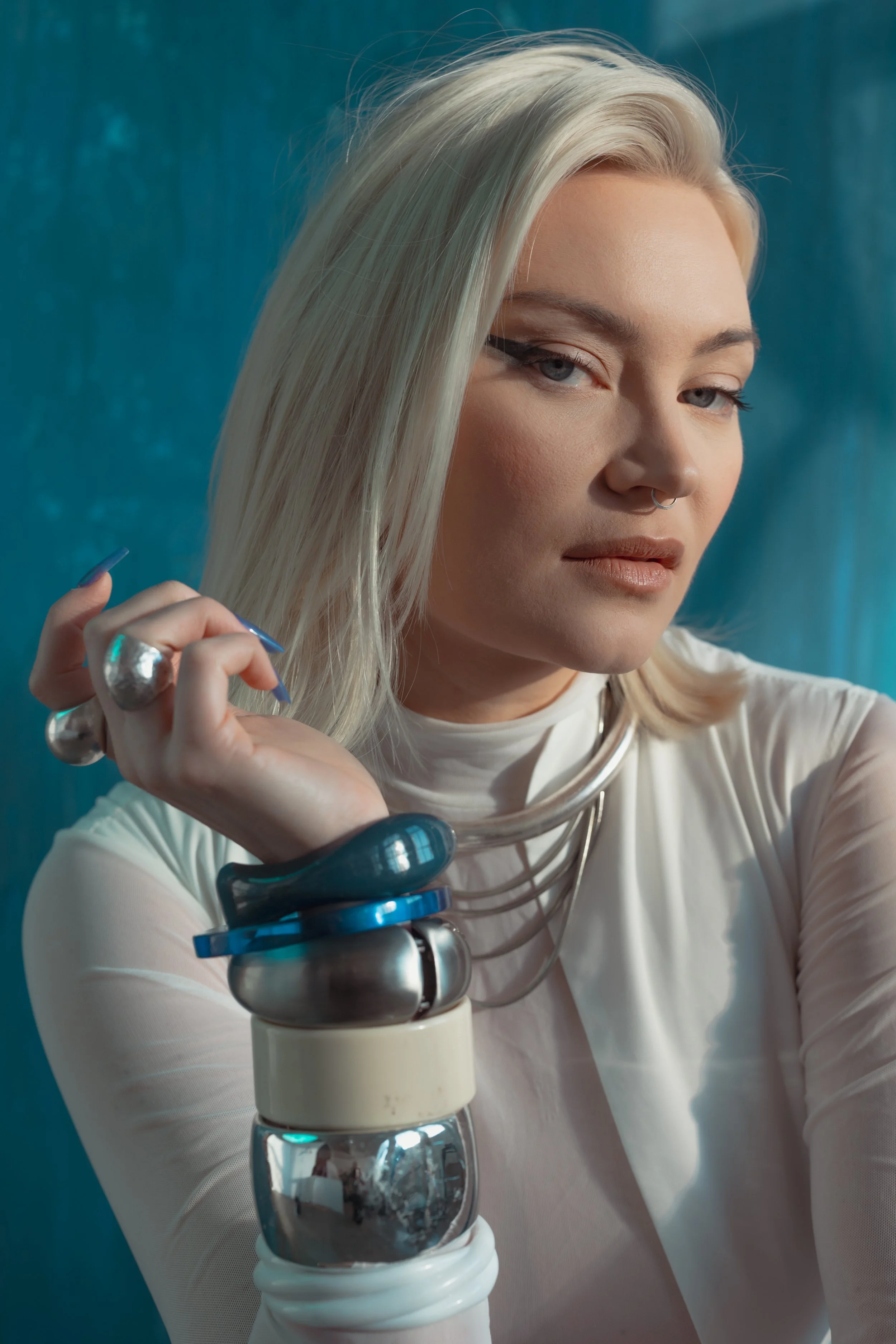 A woman with platinum blonde hair and makeup, wearing multiple rings and bracelets, is holding her hair with her left hand while looking into the camera against a blue background.