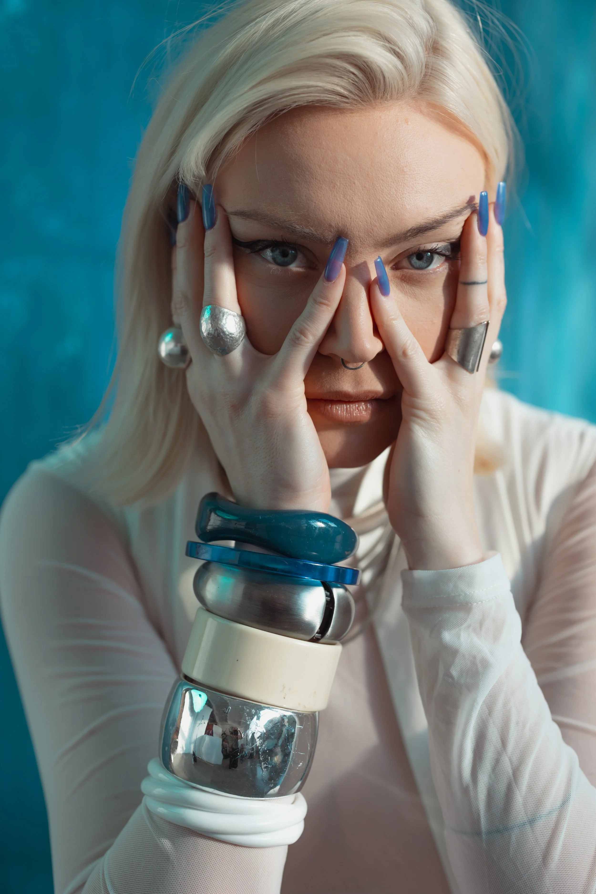 A woman with platinum blonde hair, blue eyes, and blue nail polish, wearing multiple chunky bangles and rings. She is posing with her hands around her face, looking directly at the camera against a blue background.