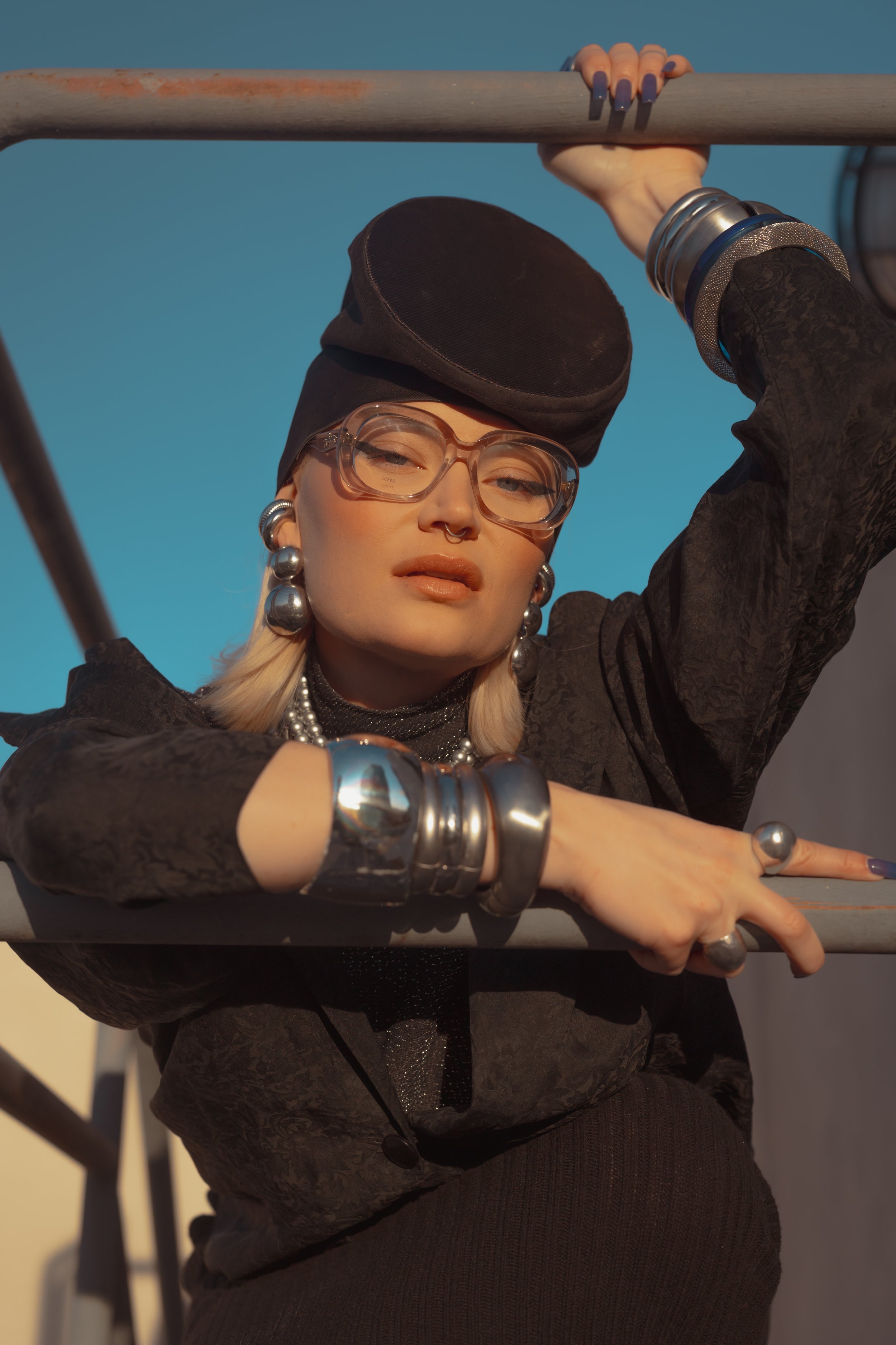 A woman holding onto a metal railing. She is wearing large silver jewelry, clear glasses, a black hat, and a dark patterned jacket, with a clear blue sky in the background.