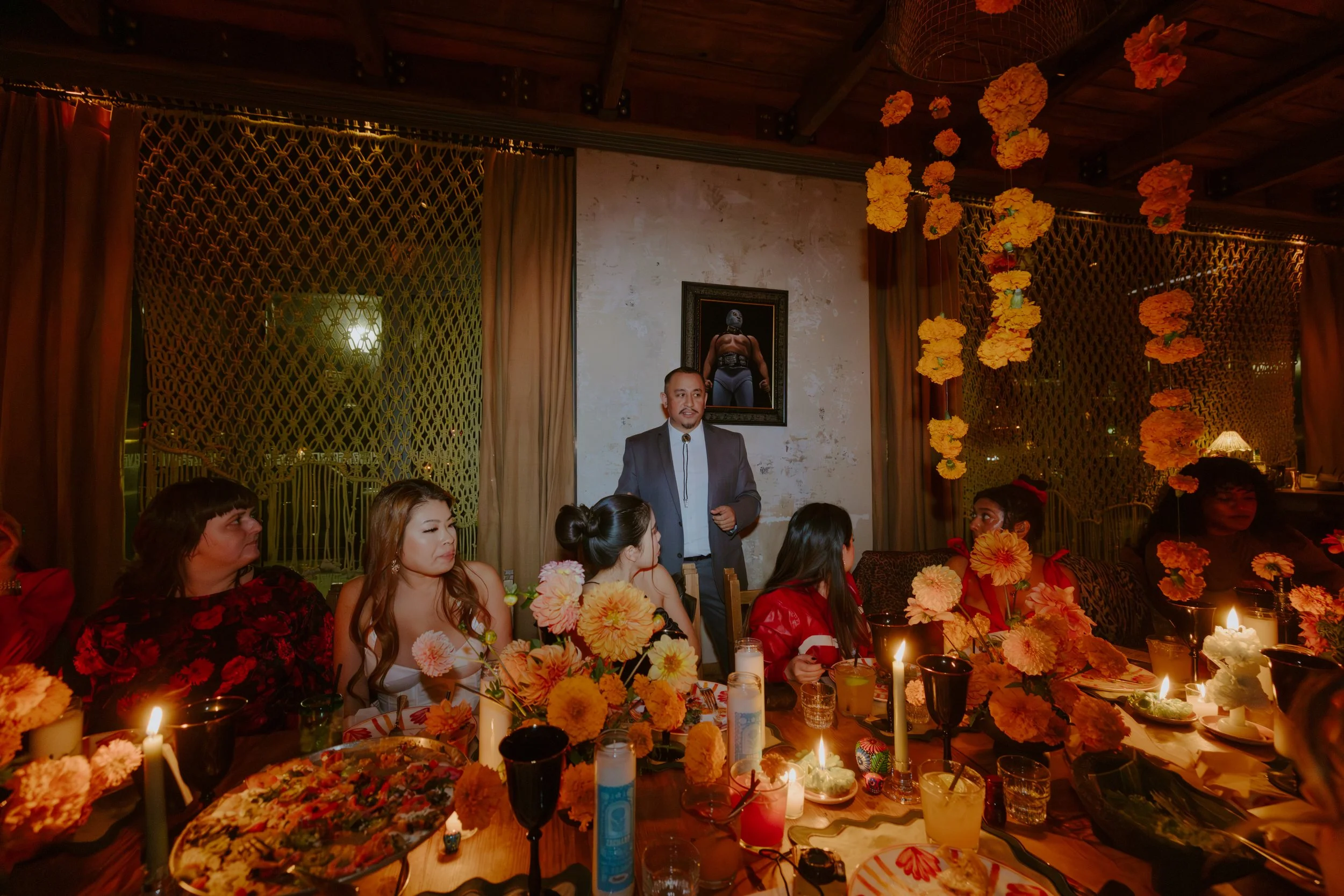 A group of women sitting around a decorated table with candles and flowers, listening to a man in a suit speaking indoors at a celebration or party.