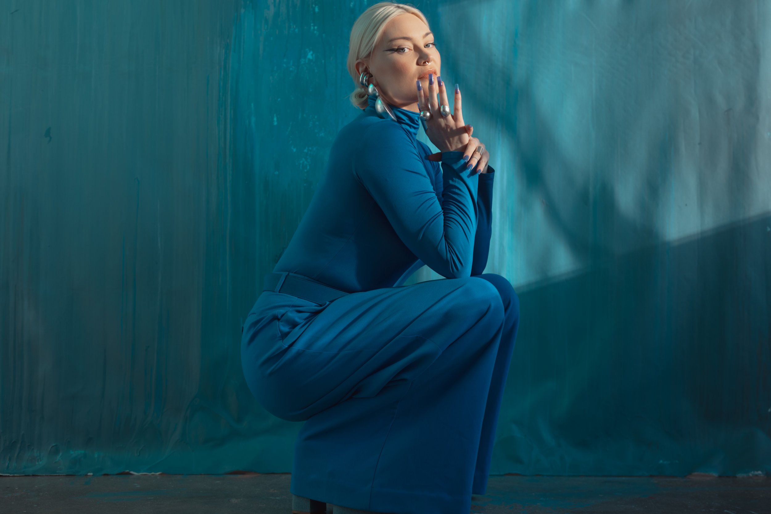 A woman with blonde hair and blue eyes wearing a blue outfit, sitting in a squatting pose against a blue-green backdrop, with her lips slightly pursed and her hands near her face, featuring large earrings and rings.
