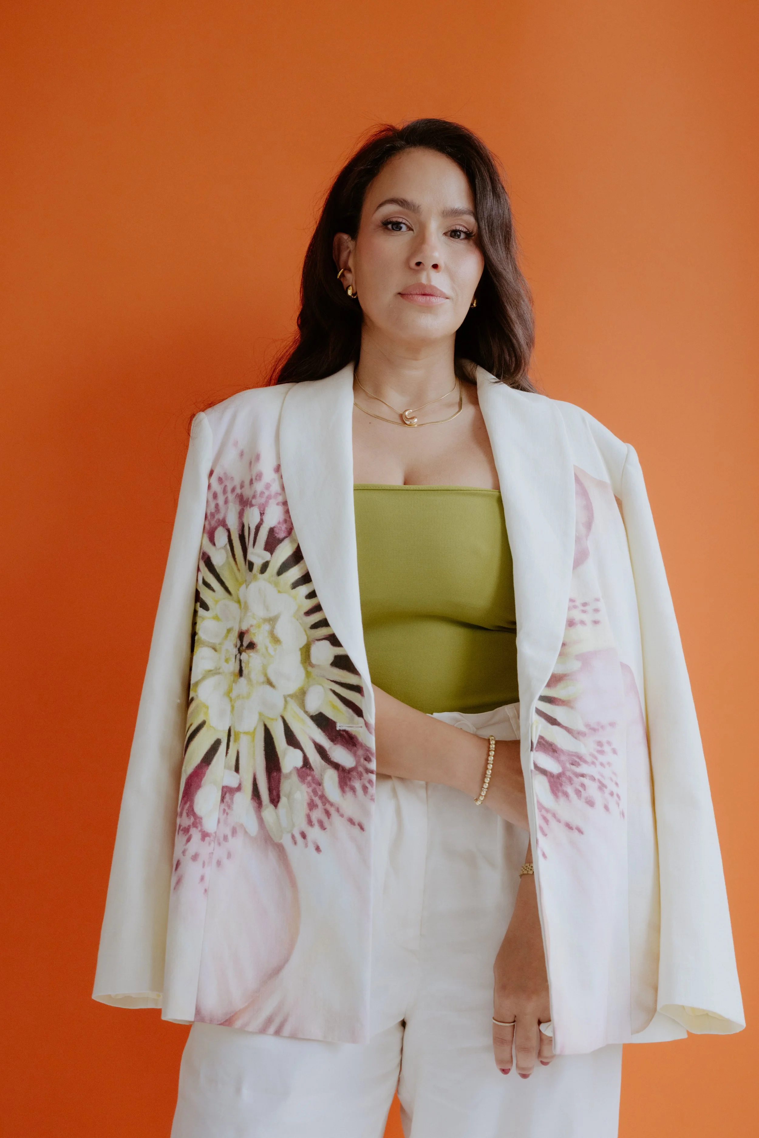 Woman wearing a green top and white pants, with a floral blazer, standing against an orange background.