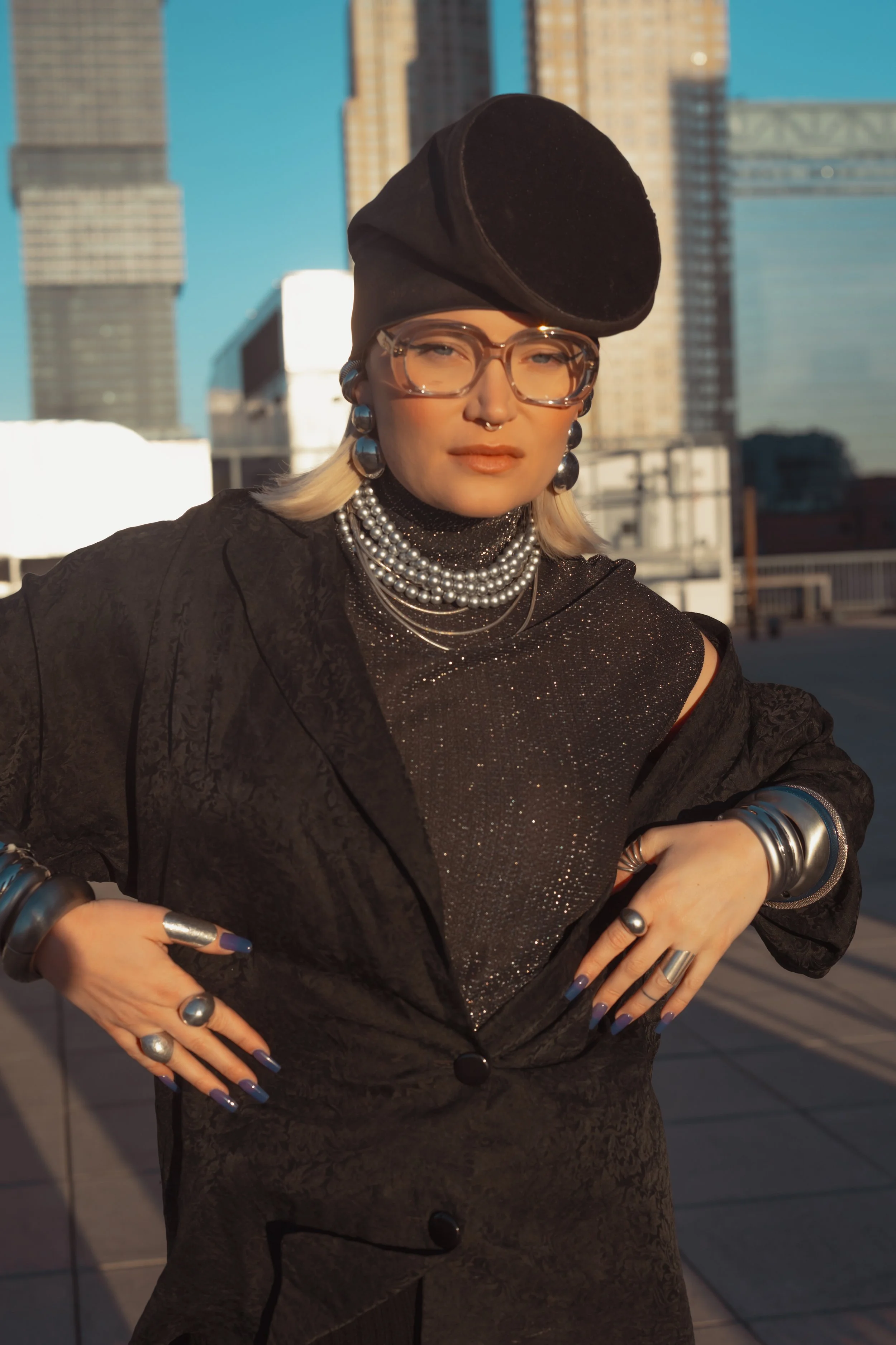 Woman in black fashion attire with jewelry, wearing glasses and a black beret, standing on a rooftop in an urban setting with tall buildings, during sunset.