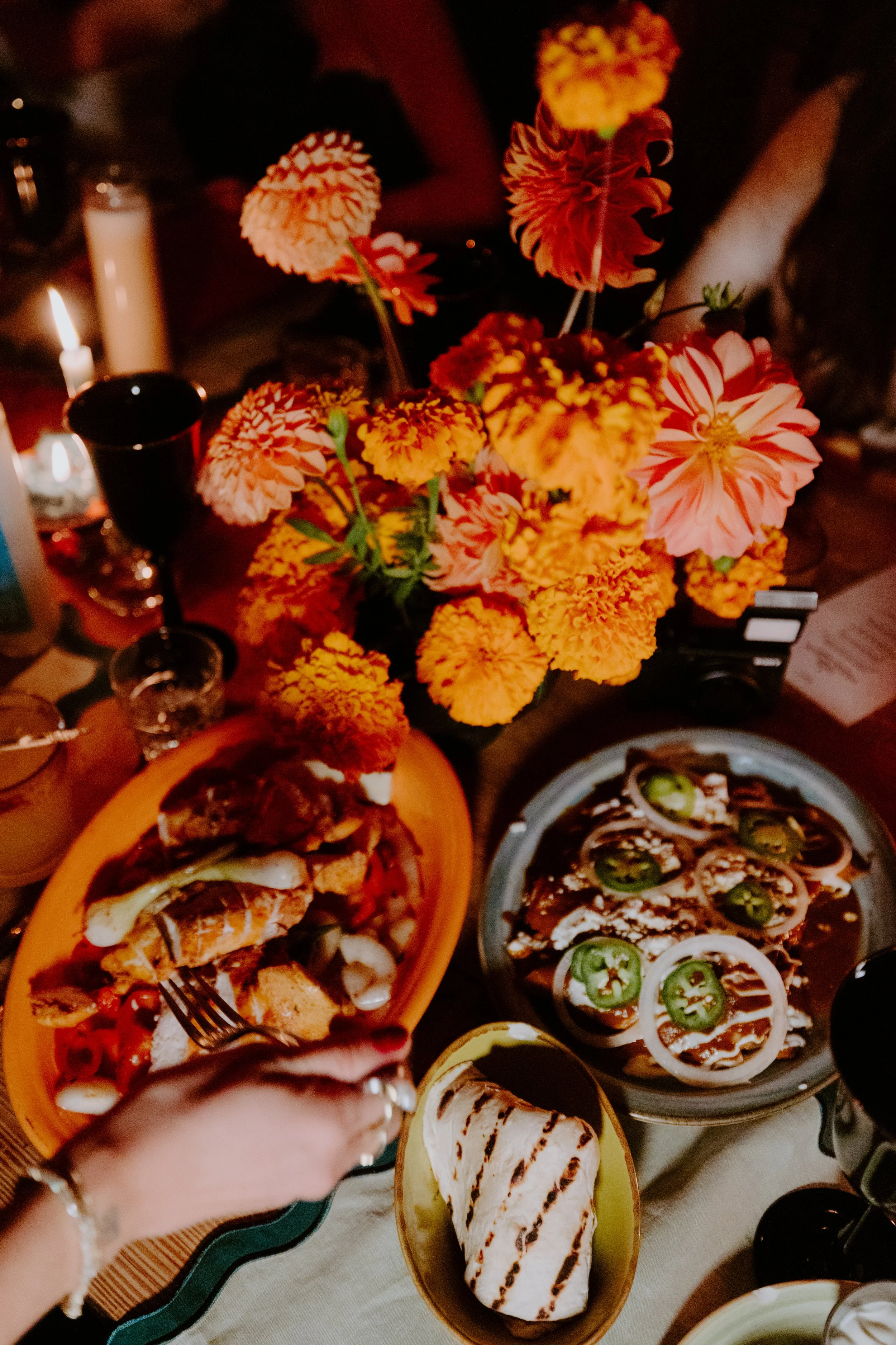 A table with various dishes, including tacos with jalapenos, grilled marshmallow, a bowl of granola, and a floral arrangement of orange and pink dahlias.