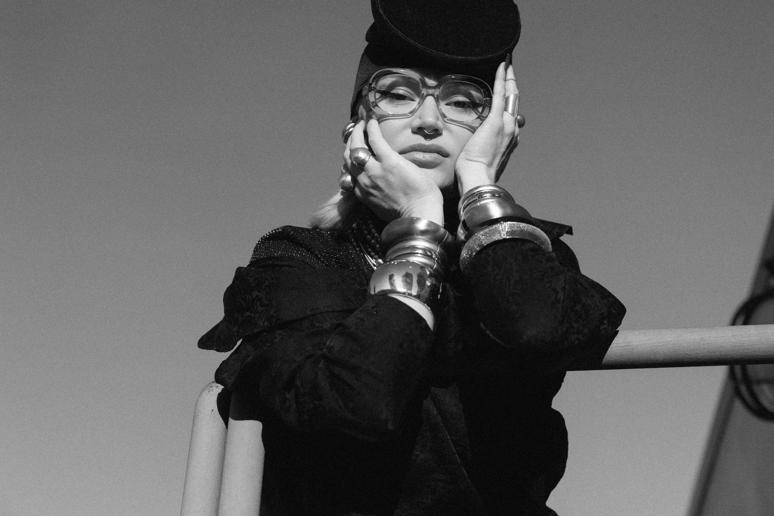 A woman with glasses, multiple bracelets, rings, and a nose ring, resting her face in her hands against a railing outdoors, with clear sky background.