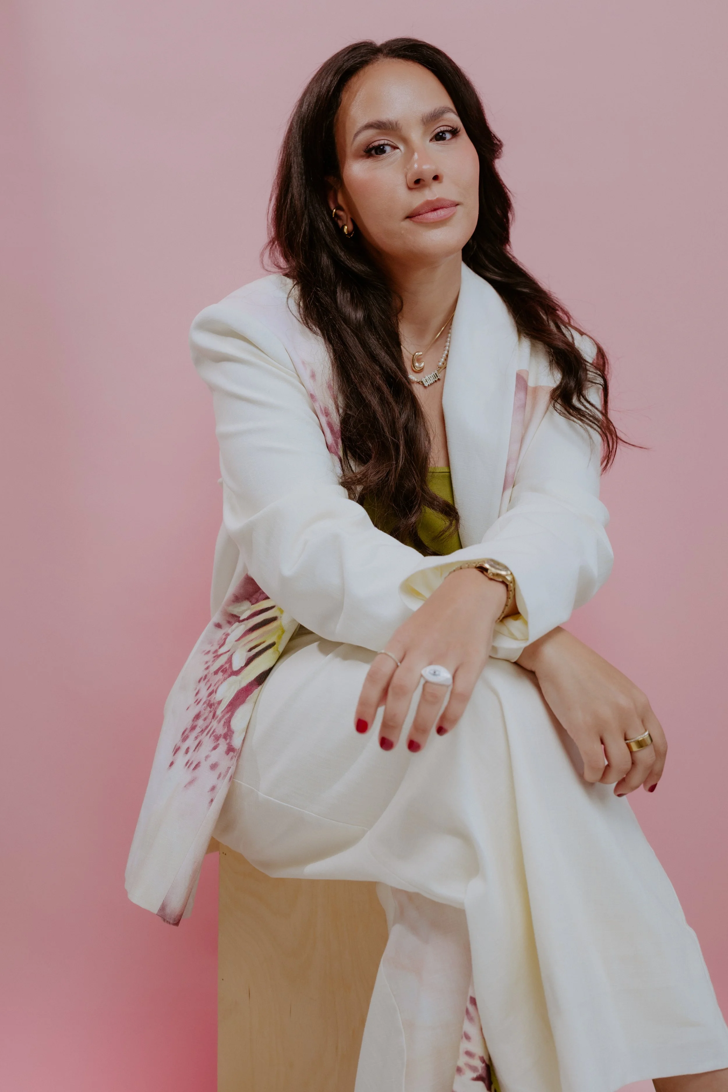 A woman with long dark hair, wearing a white blazer with pink patterns, sitting on a small wooden stool against a pink background, looking at the camera.